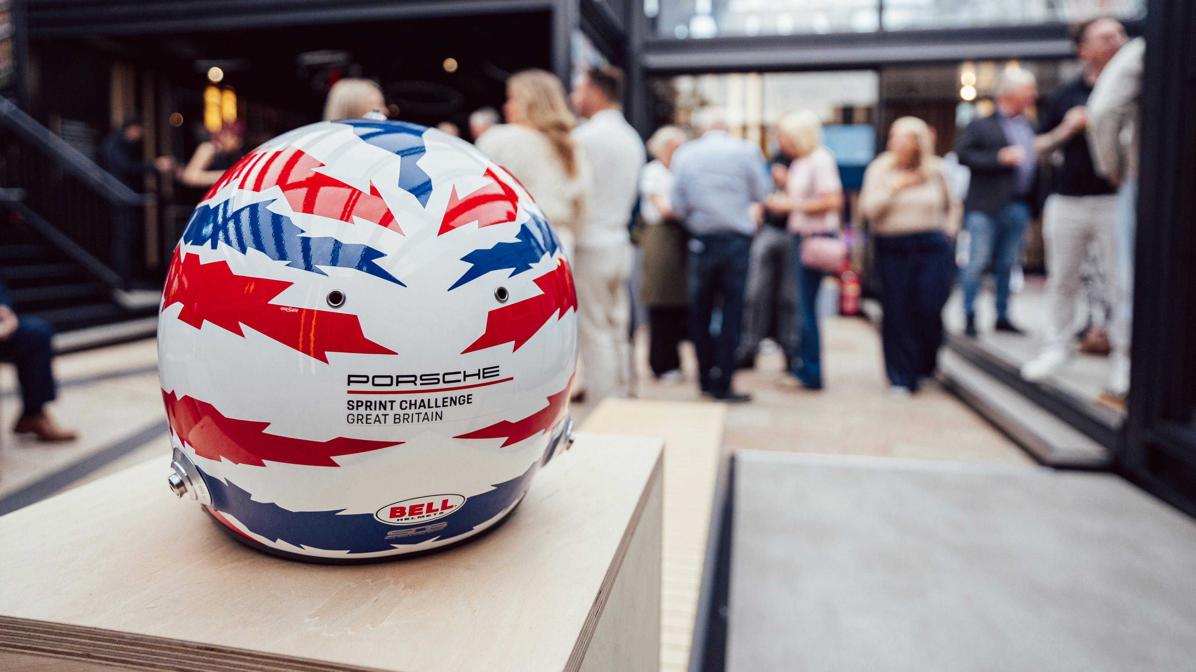 A close-up of a white racing helmet with red and blue “Union Jack” inspired lightning bolt graphics resting  on a wooden path. The text on the back read “PORSCHE SPRINT CHALLENGE GREAT BRITAIN”. In the blurred background, a group of people socialising at an outdoor event space ​