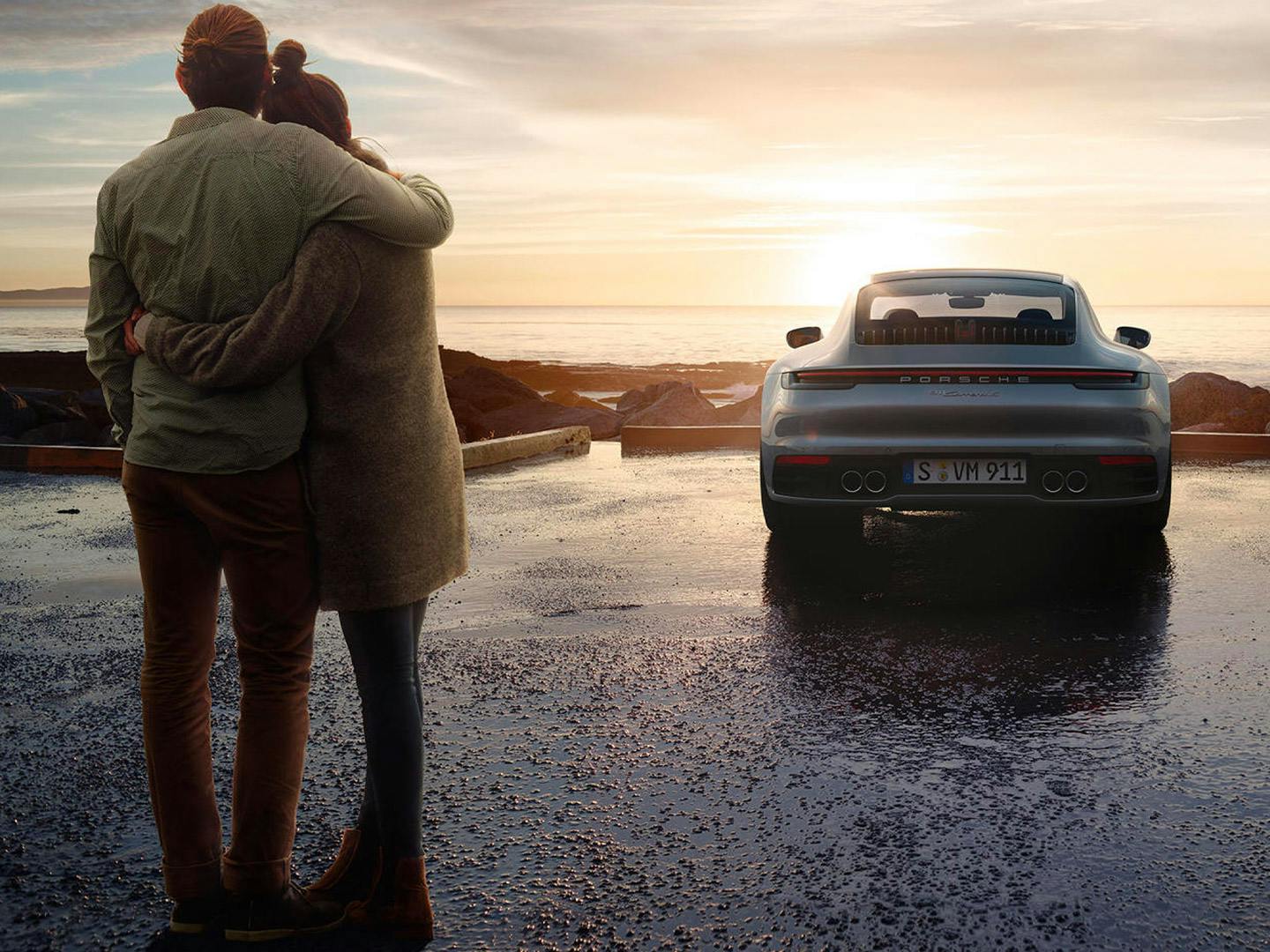 Un couple s'embrasse tendrement au coucher du soleil devant une Porsche 911, avec la mer calme en arrière-plan.