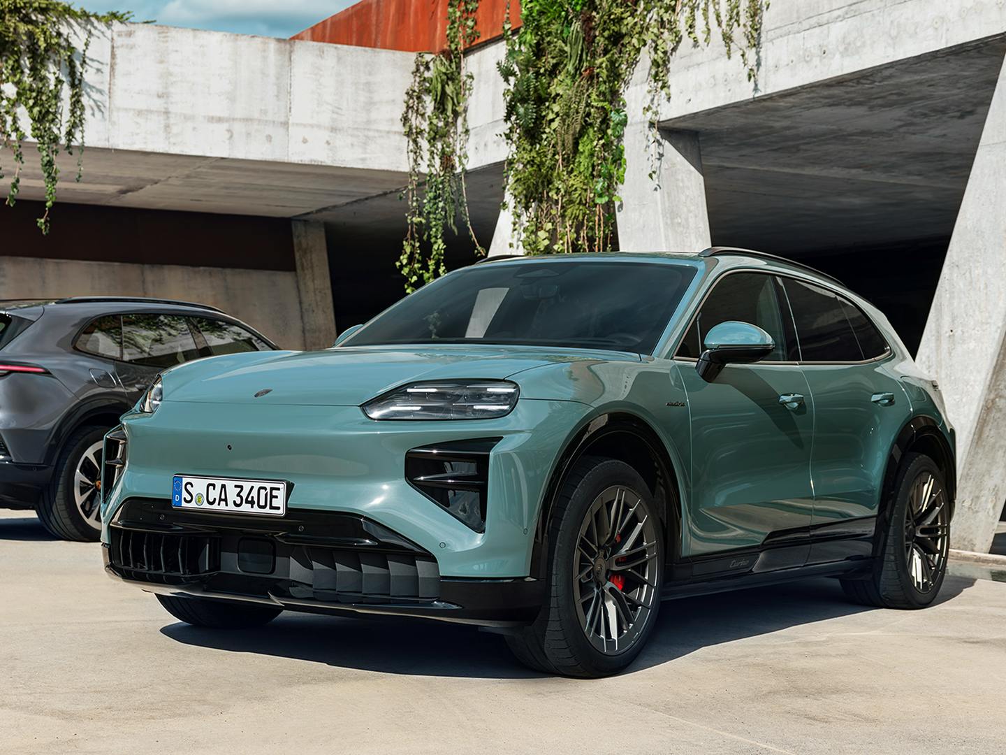 Two Porsche electric SUVs parked outside a modern concrete building with hanging greenery; a light green Porsche Cayenne Turbo Electric is in the foreground, and a dark grey Porsche Cayenne Electric is in the background.