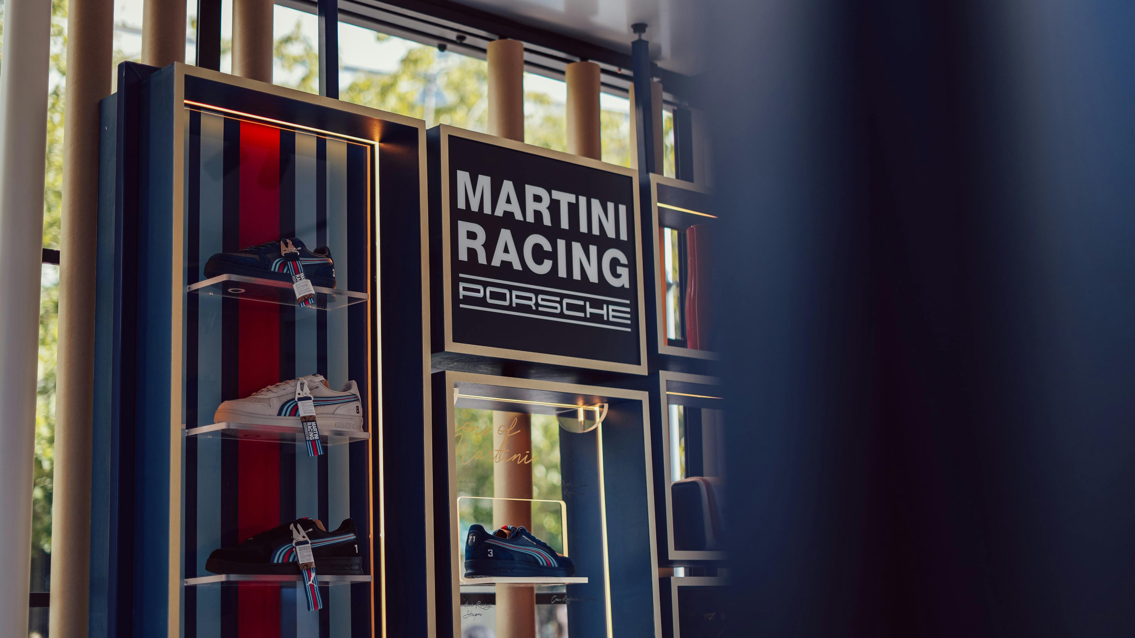 A sleek retail display for Martini Racing Porsche merchandise. On the left, three pairs of trainers (in black and white) are arranged on glass shelves against a backdrop of the iconic Martini vertical blue and red racing stripes. ​