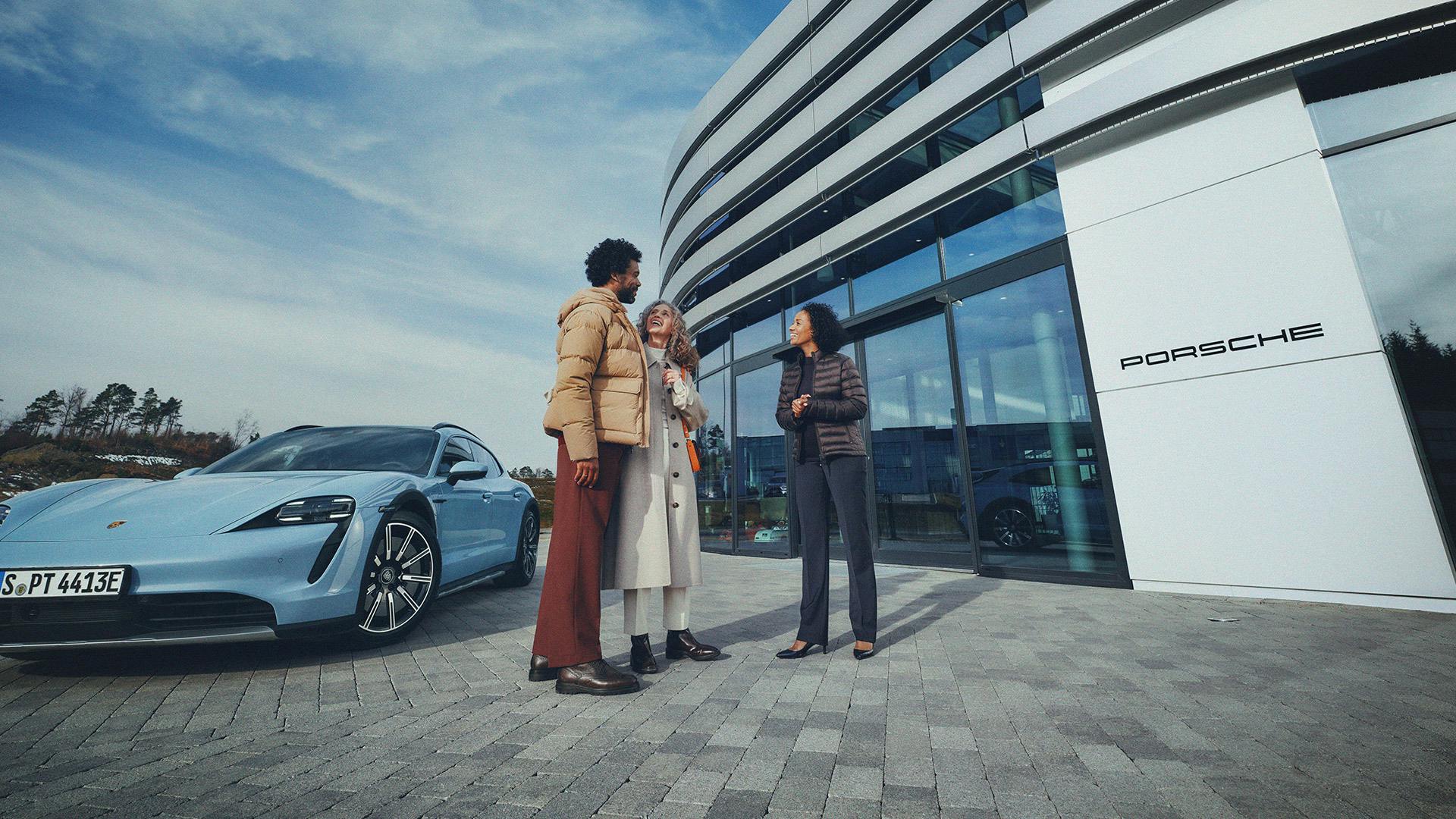 Three people standing and talking outside a modern Porsche building with large glass windows; a light blue Porsche sports car is parked nearby under a partly cloudy sky.