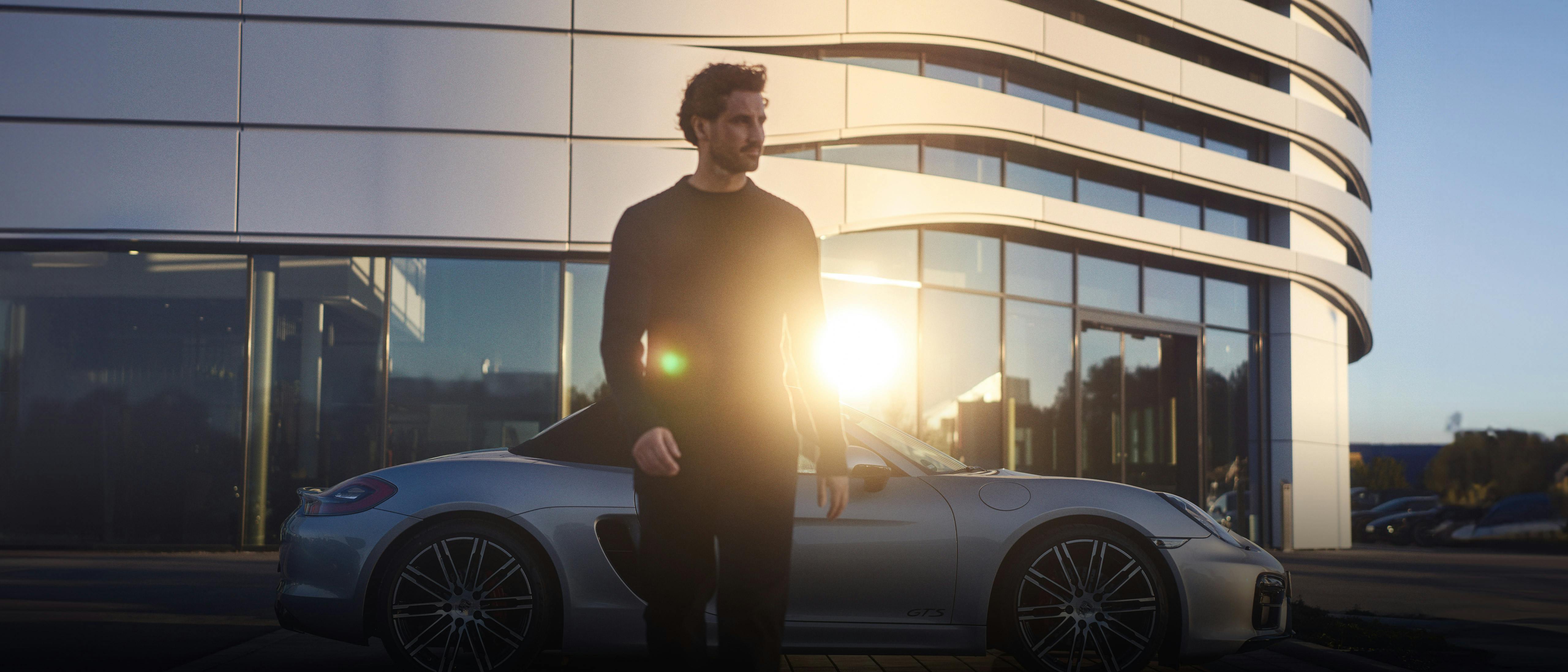 A man in a black outfit stands near a silver Porsche sports car with modern architecture and sunlight in the background.