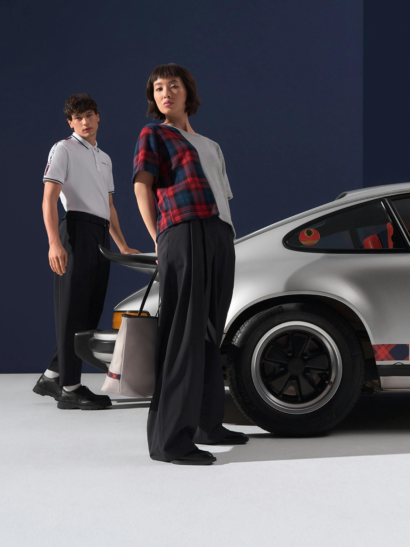 Deux jeunes personnes portant des vêtements de la collection Porsche, debout devant une Porsche 911 classique argentée.