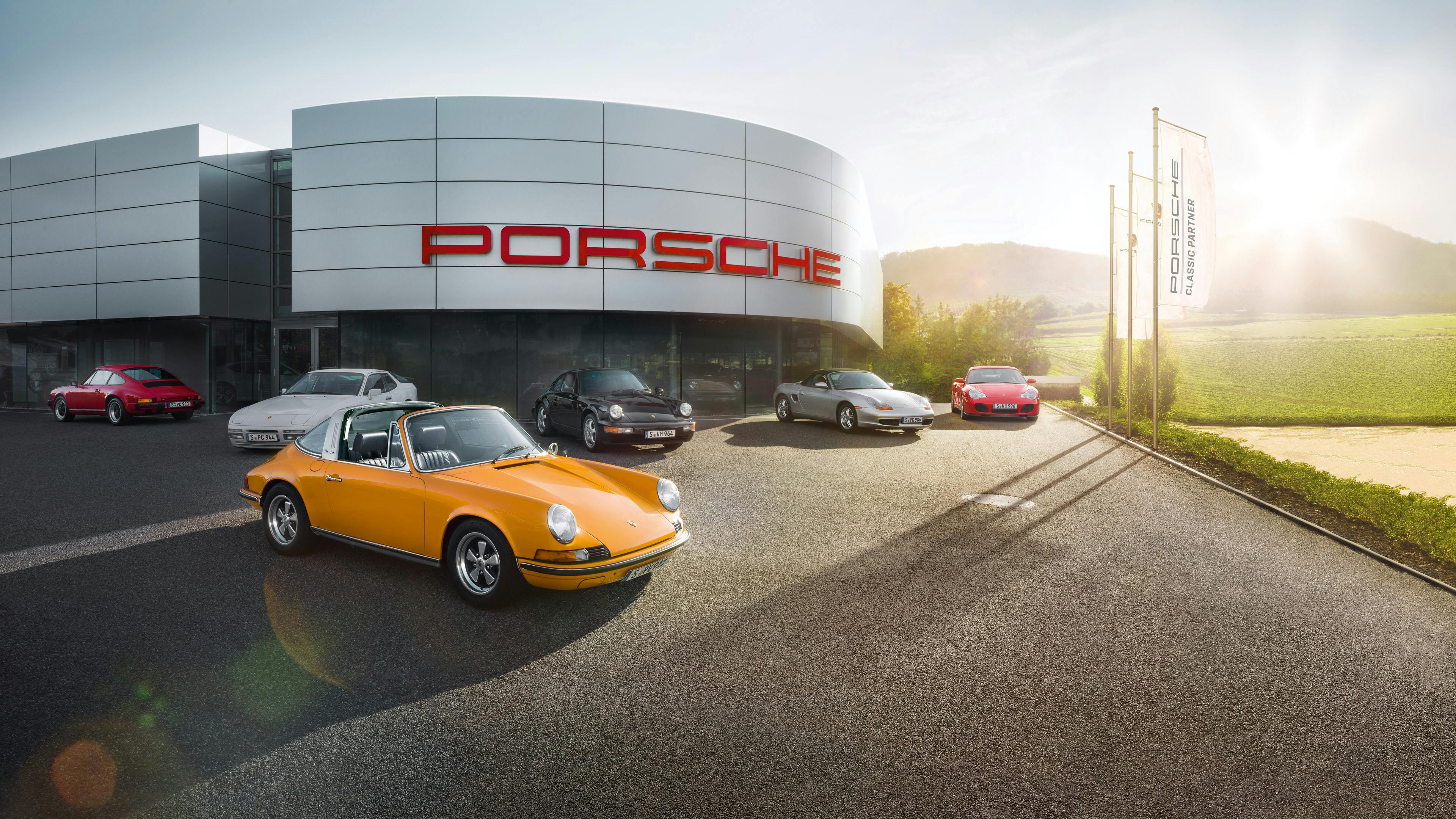 Verschiedene Porsche Modelle (vom 356 bis zum Carrera GT) auf dem Parkplatz eines Classic Partners mit gelbem 911er Carrera Cabriolet Classic im Vordergrund.