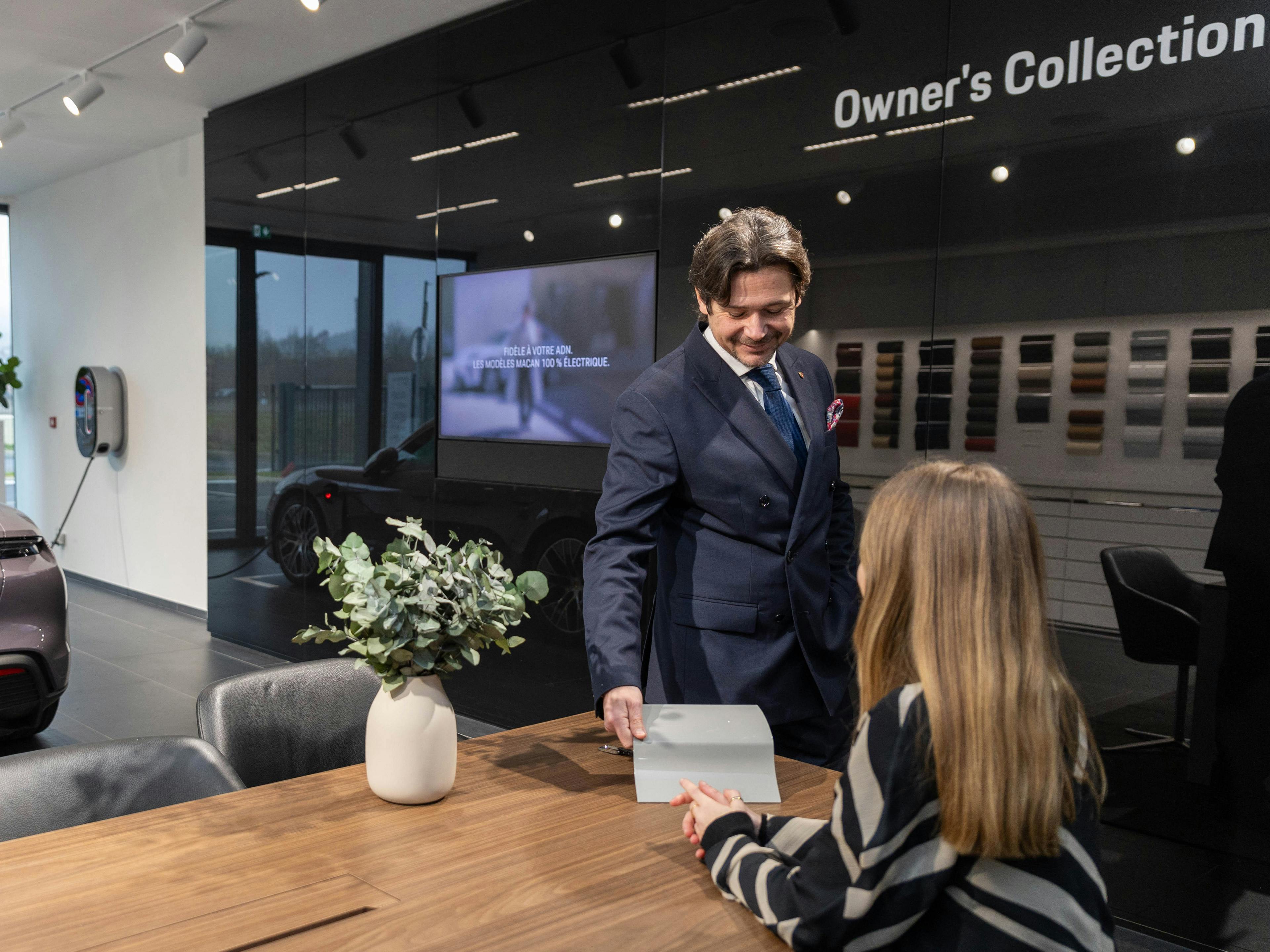 Un collaborateur de Porsche debout à côté d'un bureau dans une salle d'exposition montre un exemple de peinture de voiture gris clair à une cliente assise au bureau.