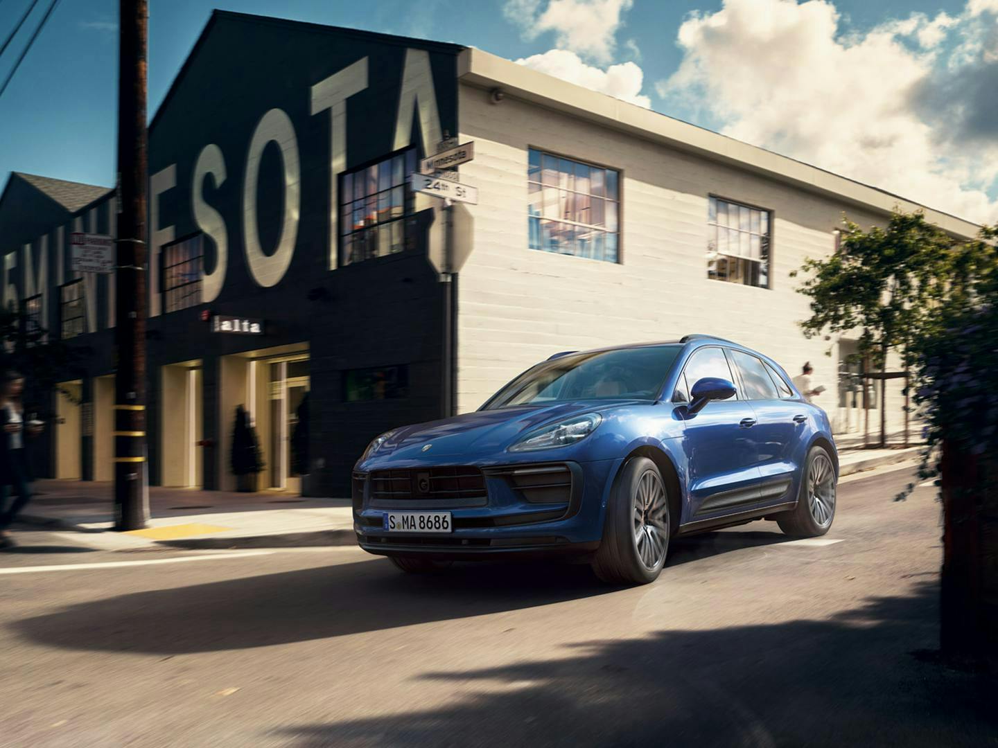 The Macan driving along a road, going past a building that a white wall on one side and a brown wall on the front.