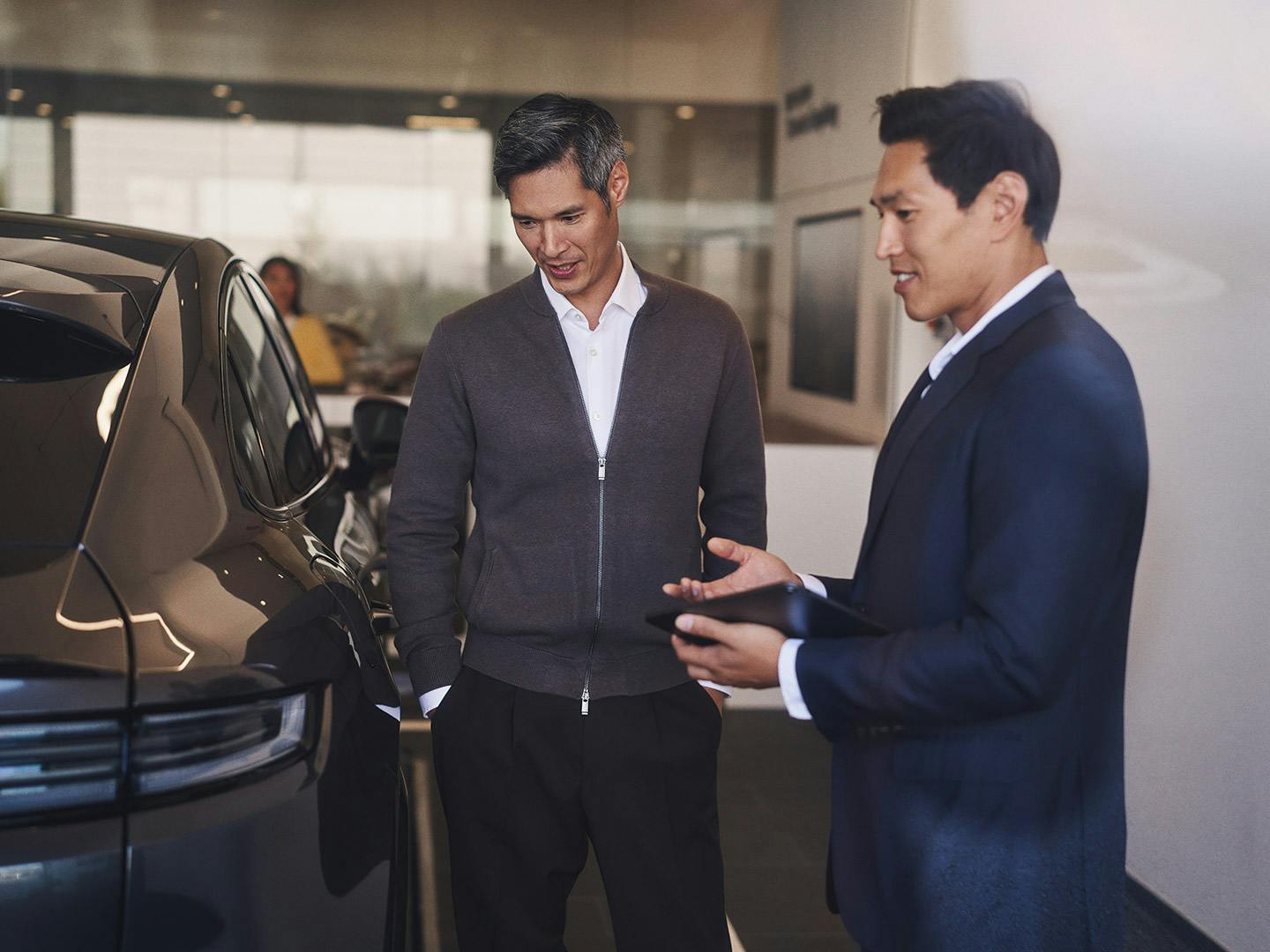 Porsche employee in a blue suit presents a Porsche car to a couple in the showroom.
