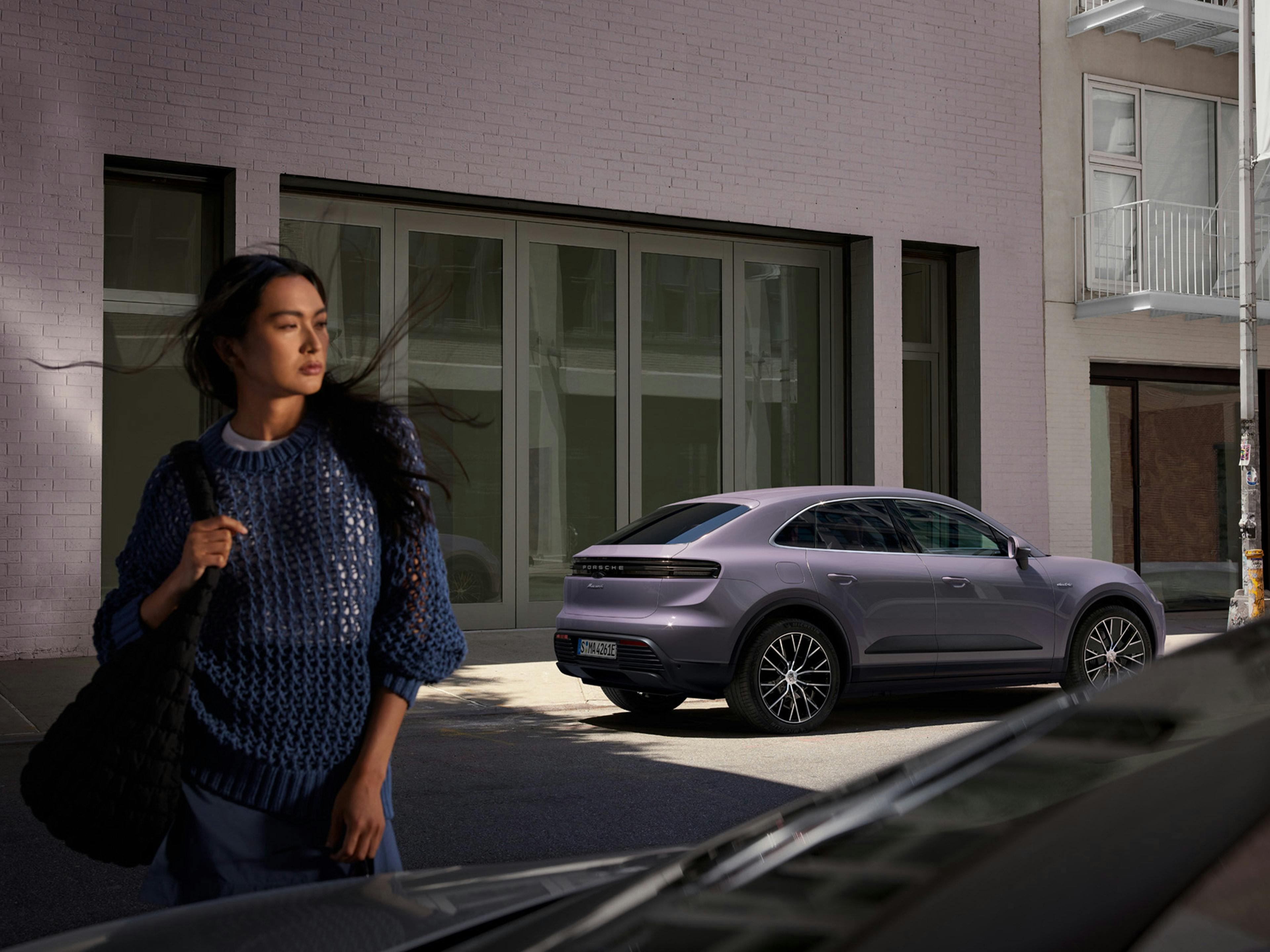 Eine junge Frau steht vor dem Porsche Macan electric in der Innenstadt.