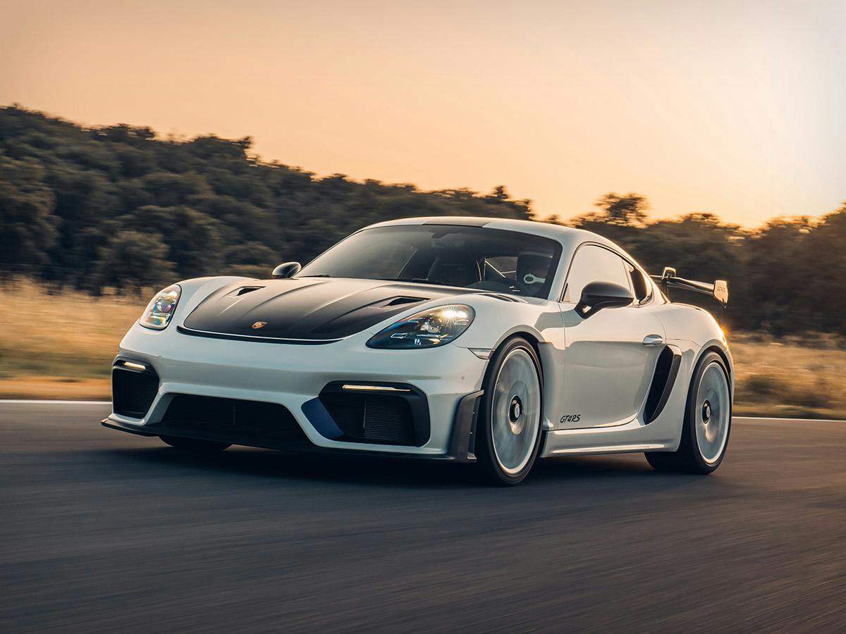 Front three‑quarter rolling shot of the 718 Cayman GT4 RS in Ziskiwhite with Violet tinted carbon, driving on a track at sunset.