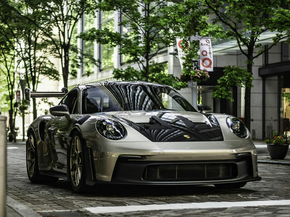 Front three-quarter view of a Porsche 911 GT3 RS in Paint to Sample colour Palladiummetallic, parked on a cobblestone street with trees and buildings in the background.