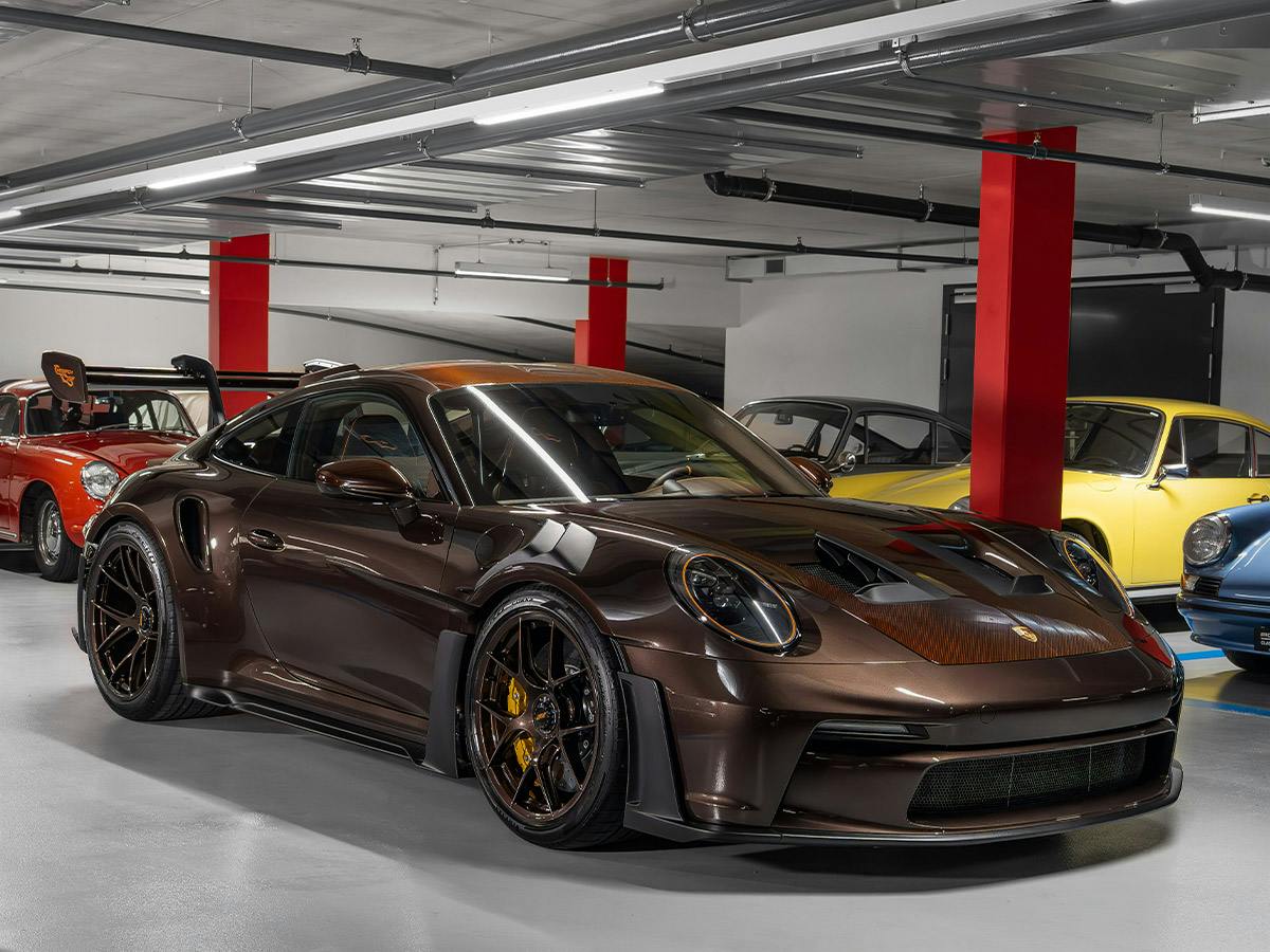 911 GT3 RS in exclusive Paint to Sample colour Macadamiabrown with brown-tinted carbon roof and bonnet, shown from a front-side angle in a garage alongside several classic and modern sports cars.