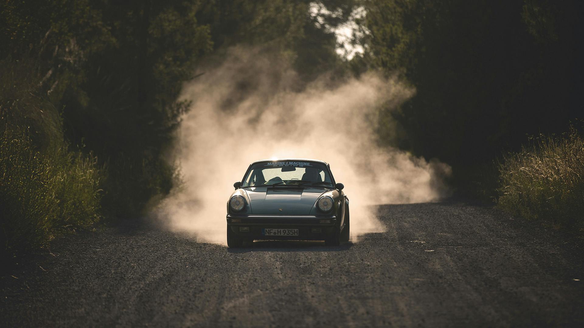 A Porsche 911 on a gravel road, surrounded by swirling dust.