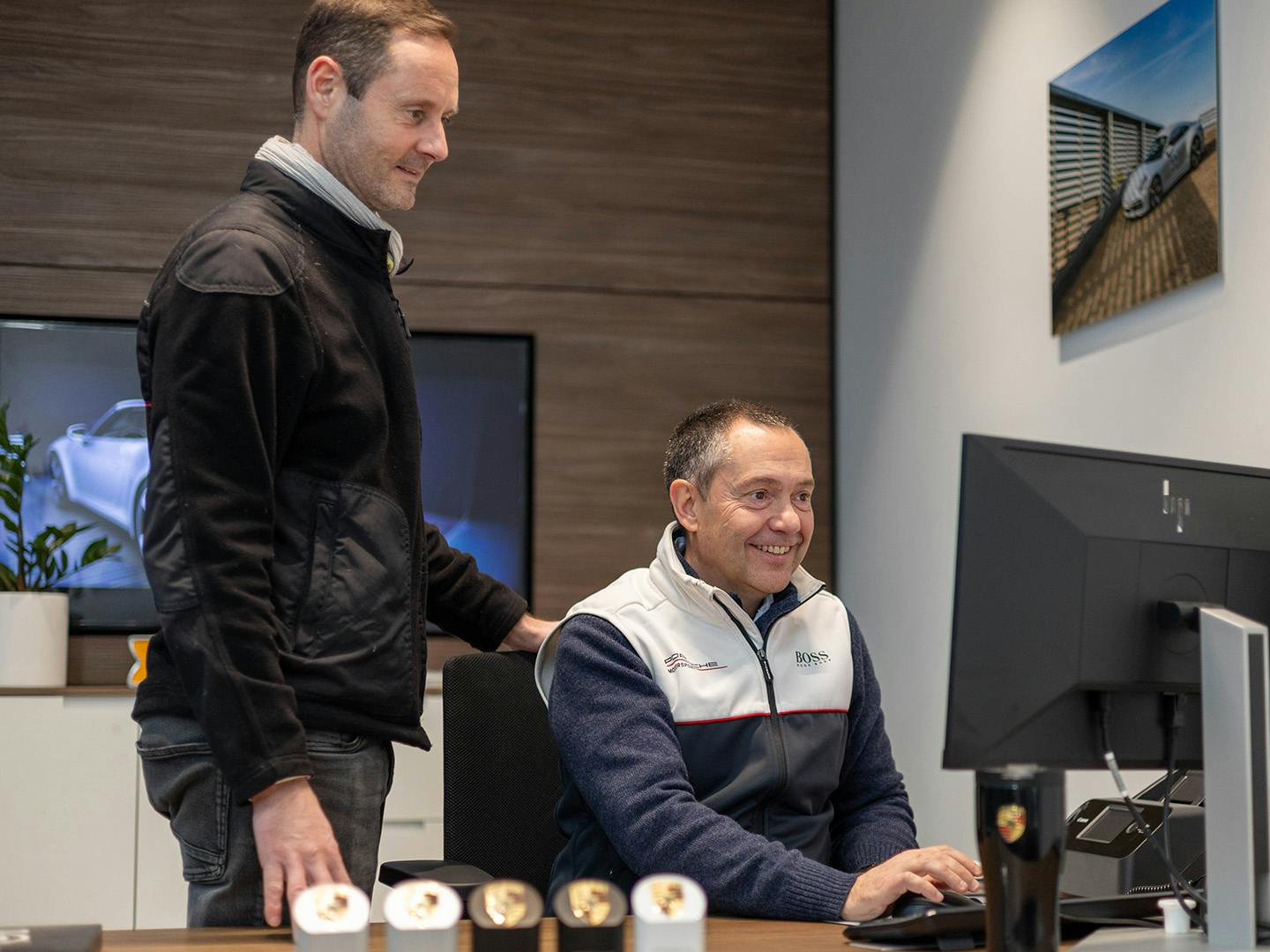 Un employé de Porsche, assis derrière un bureau, tape sur le clavier d'un ordinateur tandis qu'un collègue se tient à côté de lui et regarde l'écran de l'ordinateur.