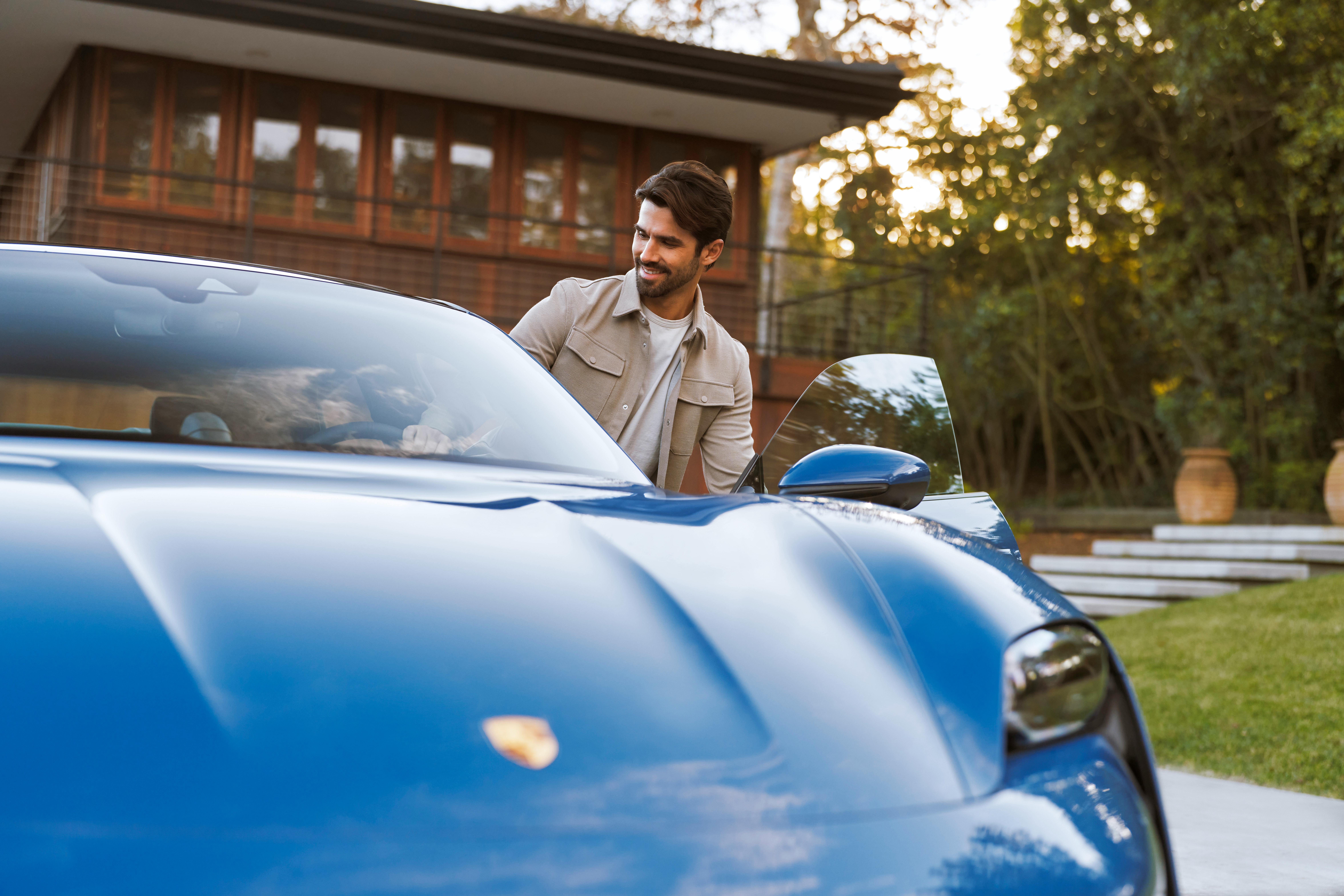 A sleek blue Porsche parked outdoors near a modern house, with a person standing beside the open car door amidst greenery and evening light.