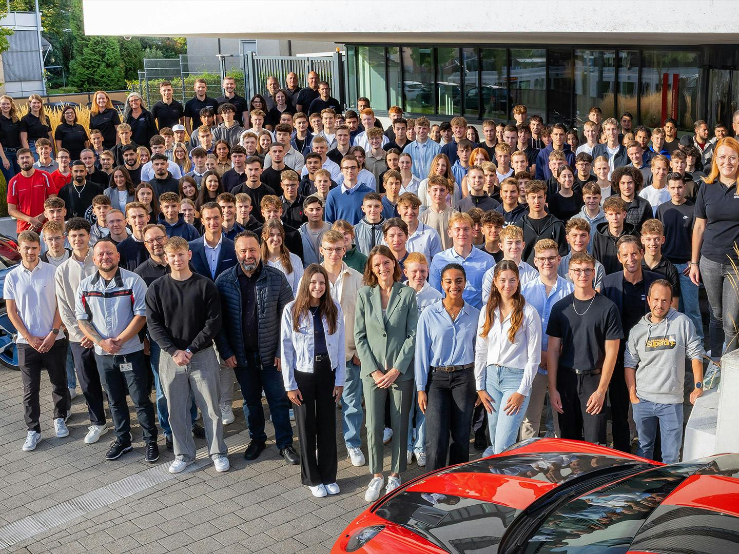 Große Gruppe von Porsche Auszubildenden und Porsche Mitarbeitenden vor modernem Firmengebäude; im Vordergrund zwei Fahrzeuge mit Ausbildungsbranding.