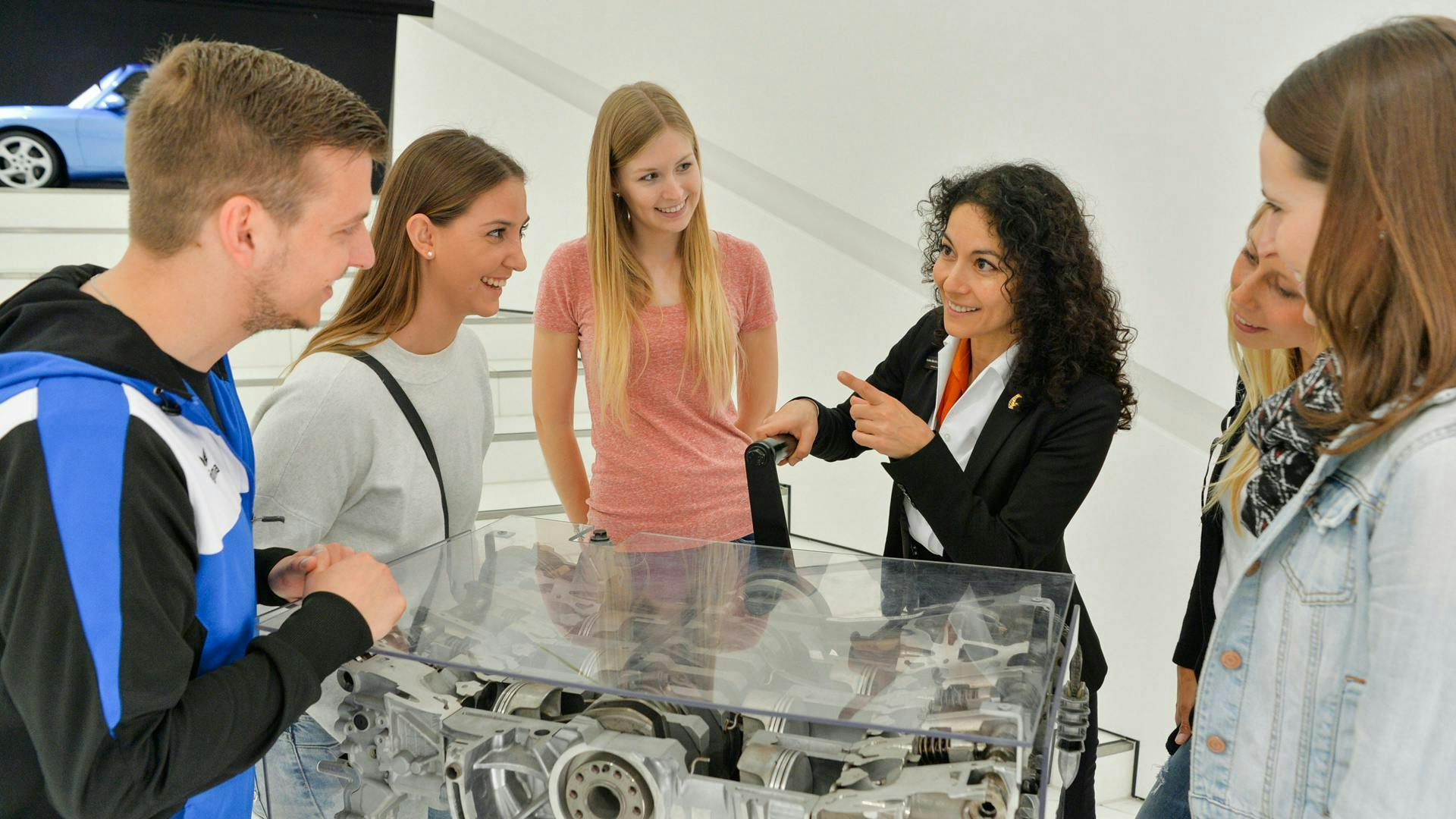 Young people standing around an exhibited Porsche engine and smiling while listening to a museum guide