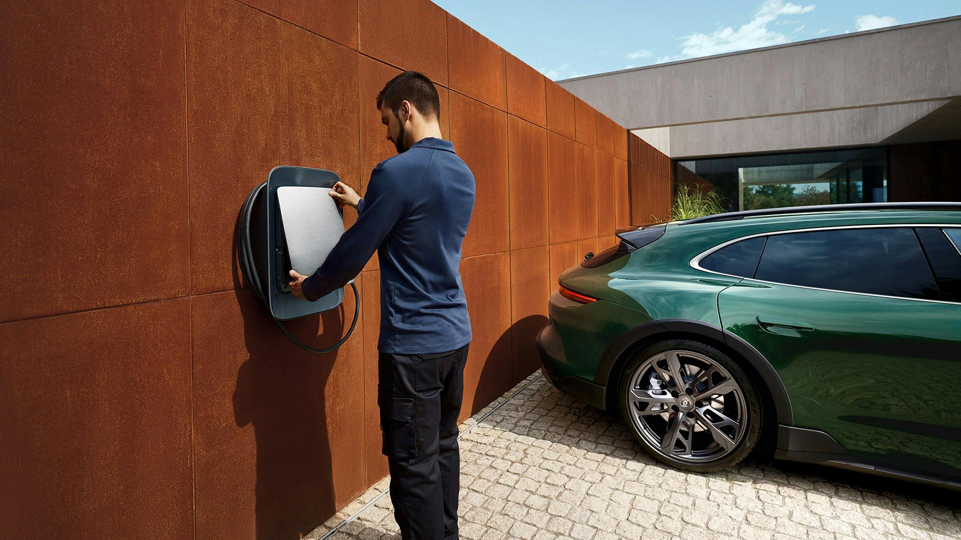Un homme branche une Porsche Taycan verte à un chargeur mural devant une maison contemporaine aux murs couleur rouille.