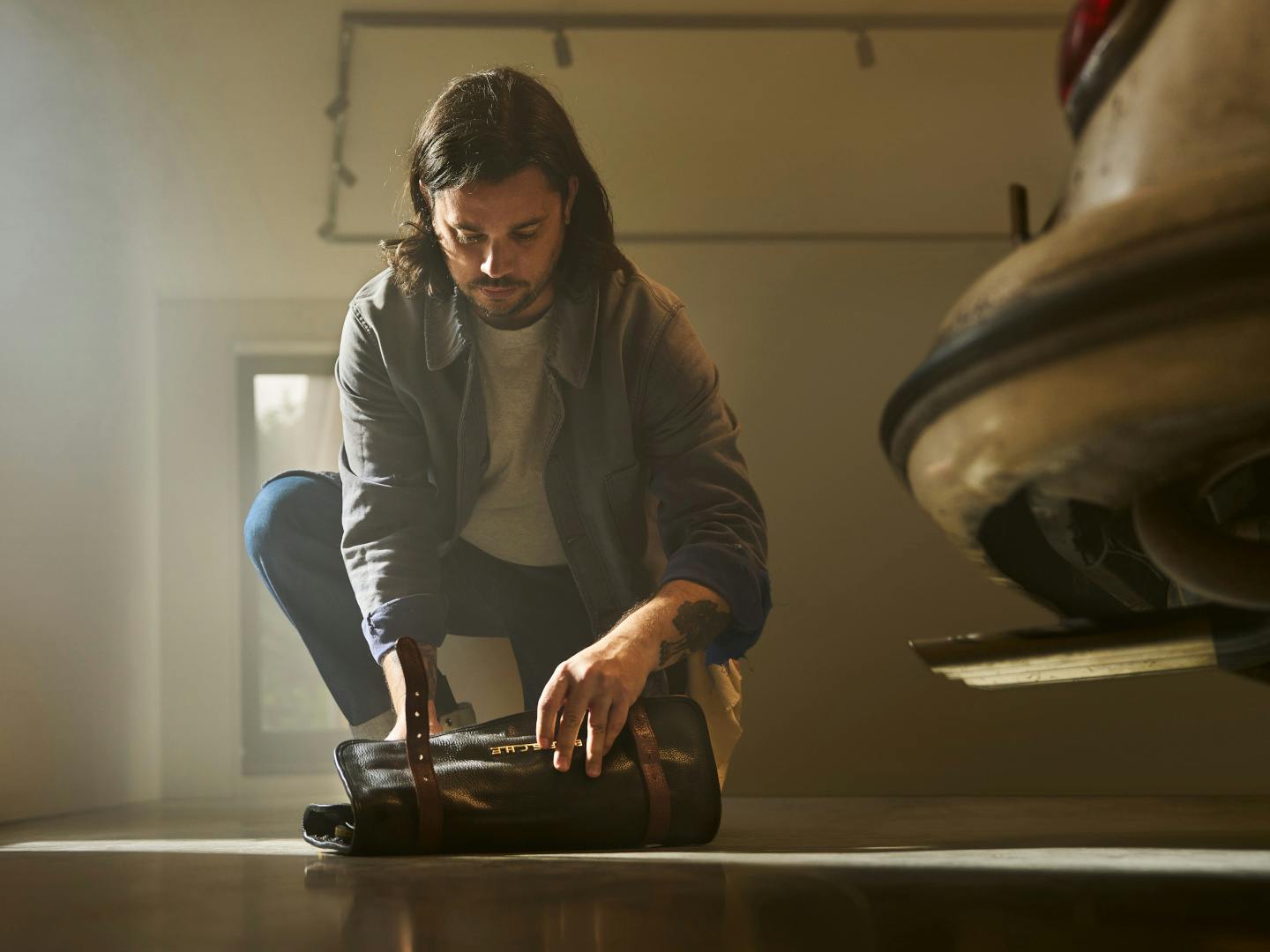 A medium shot of a man with long, dark hair and a moustache, crouching on the concrete floor of a brightly lit garage. He wears a grey button-up overshirt over a light t-shirt and blue jeans, and he is looking down while unrolling a black leather tool bag with brown leather straps. To his right, the rear bumper and exhaust pipe of a vintage car are partially visible.