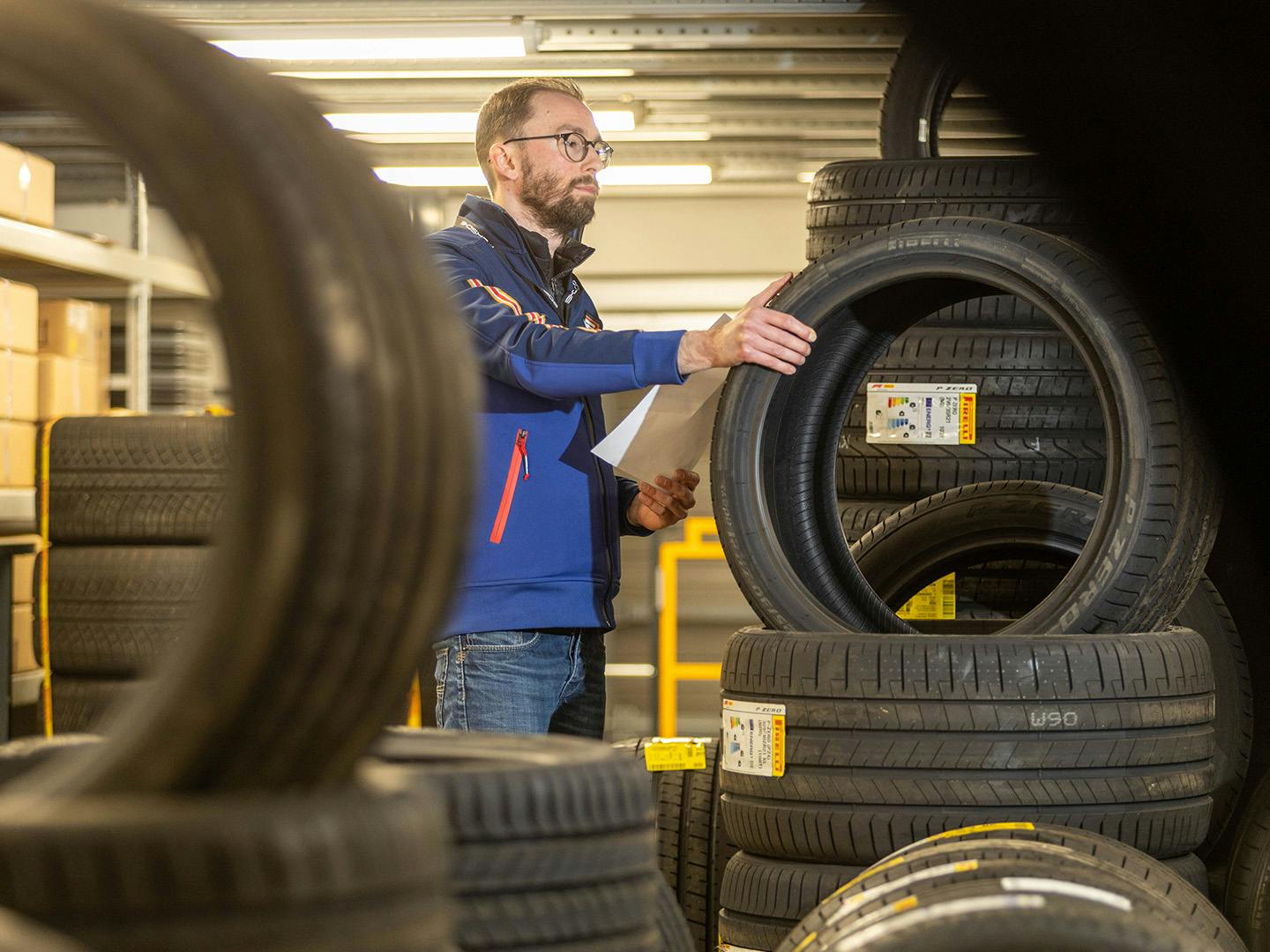 Un employé de Porsche se tient à côté d'une pile de pneus, tenant le pneu au-dessus de la pile d'une main tout en tenant un morceau de papier dans son autre main.
