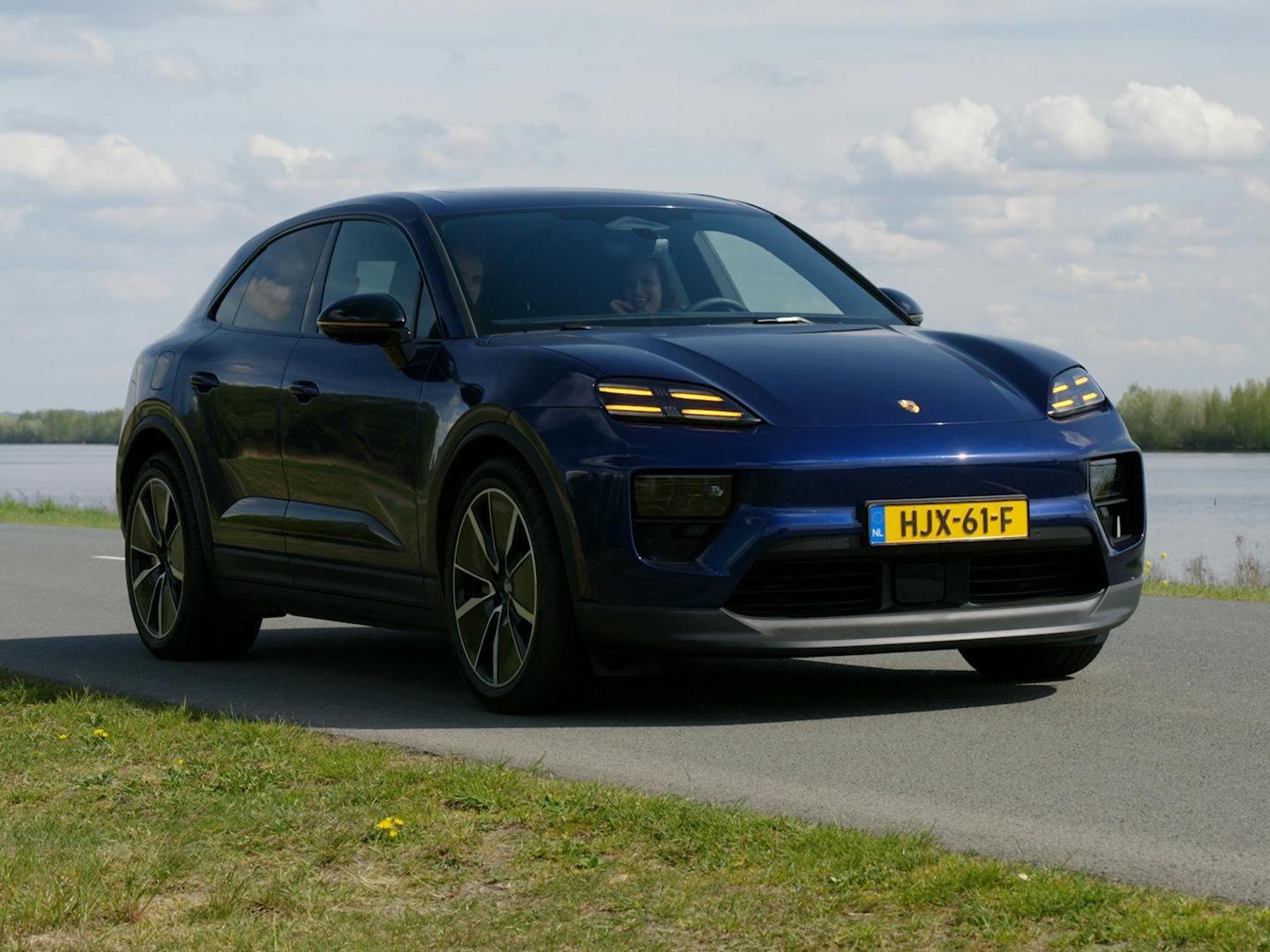 Vooraanzicht van een blauwe Porsche Macan EV die over een weg langs het water rijdt. De afbeelding benadrukt de kenmerkende vierpunts led-koplampen, de aerodynamische neus en de grote lichtmetalen velgen van deze elektrische Porsche Macan.