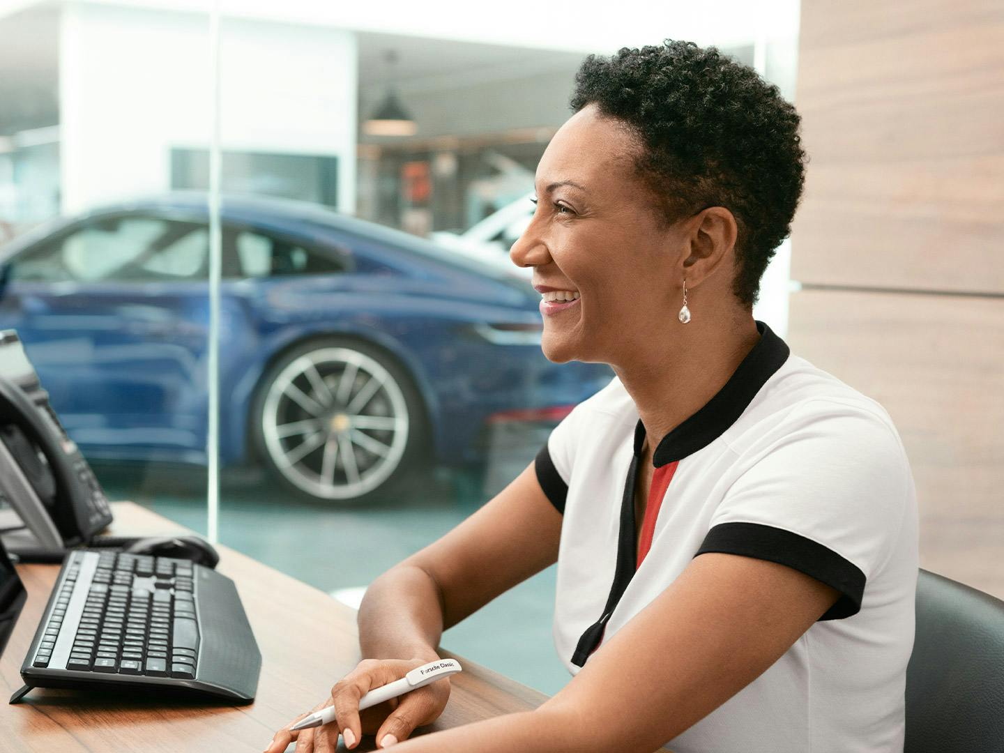 Smiling woman in a white Porsche outfit sitting at a counter in a Porsche Centre, with a blue Porsche Taycan visible in the background.