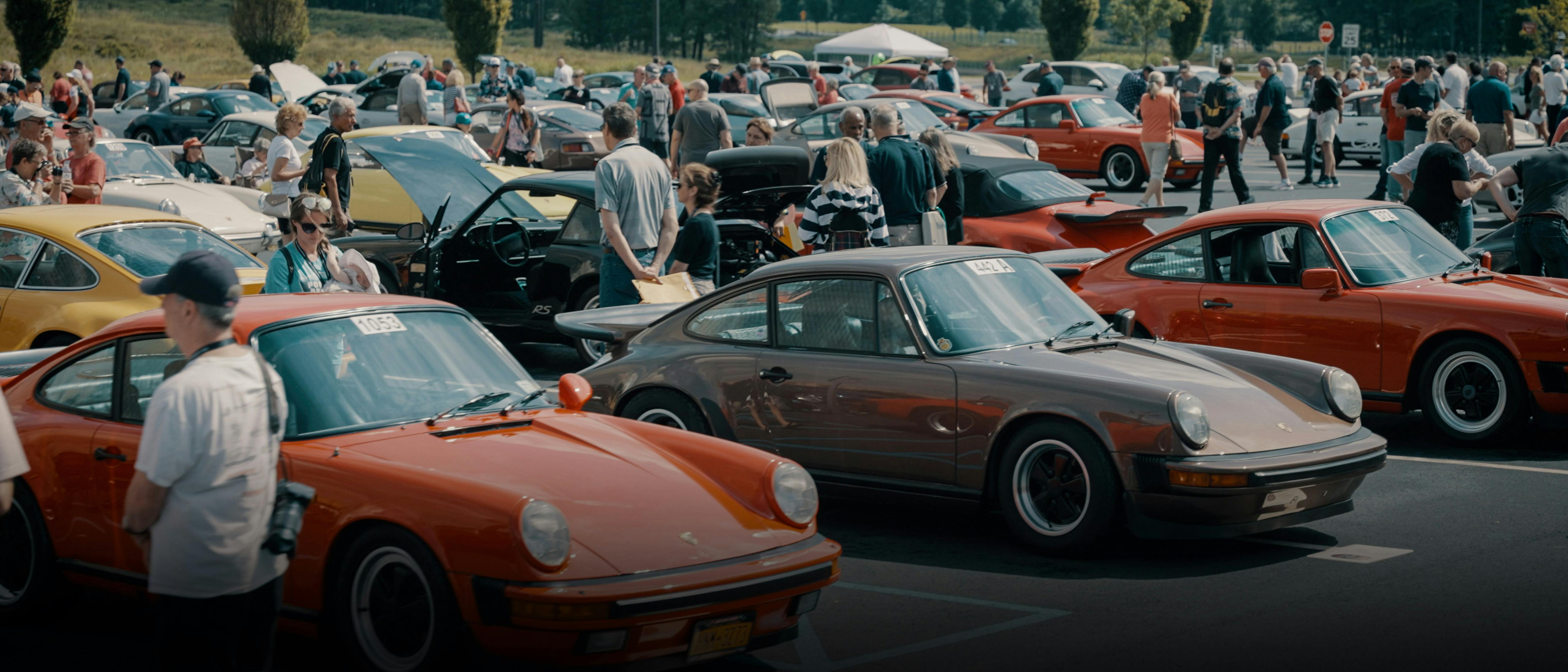 Großes Treffen klassischer Porsche‑Sportwagen auf einem Parkplatz, mit mehreren historischen Modellen in verschiedenen Farben, umgeben von zahlreichen Besucherinnen und Besuchern, die die Fahrzeuge betrachten und miteinander ins Gespräch kommen.