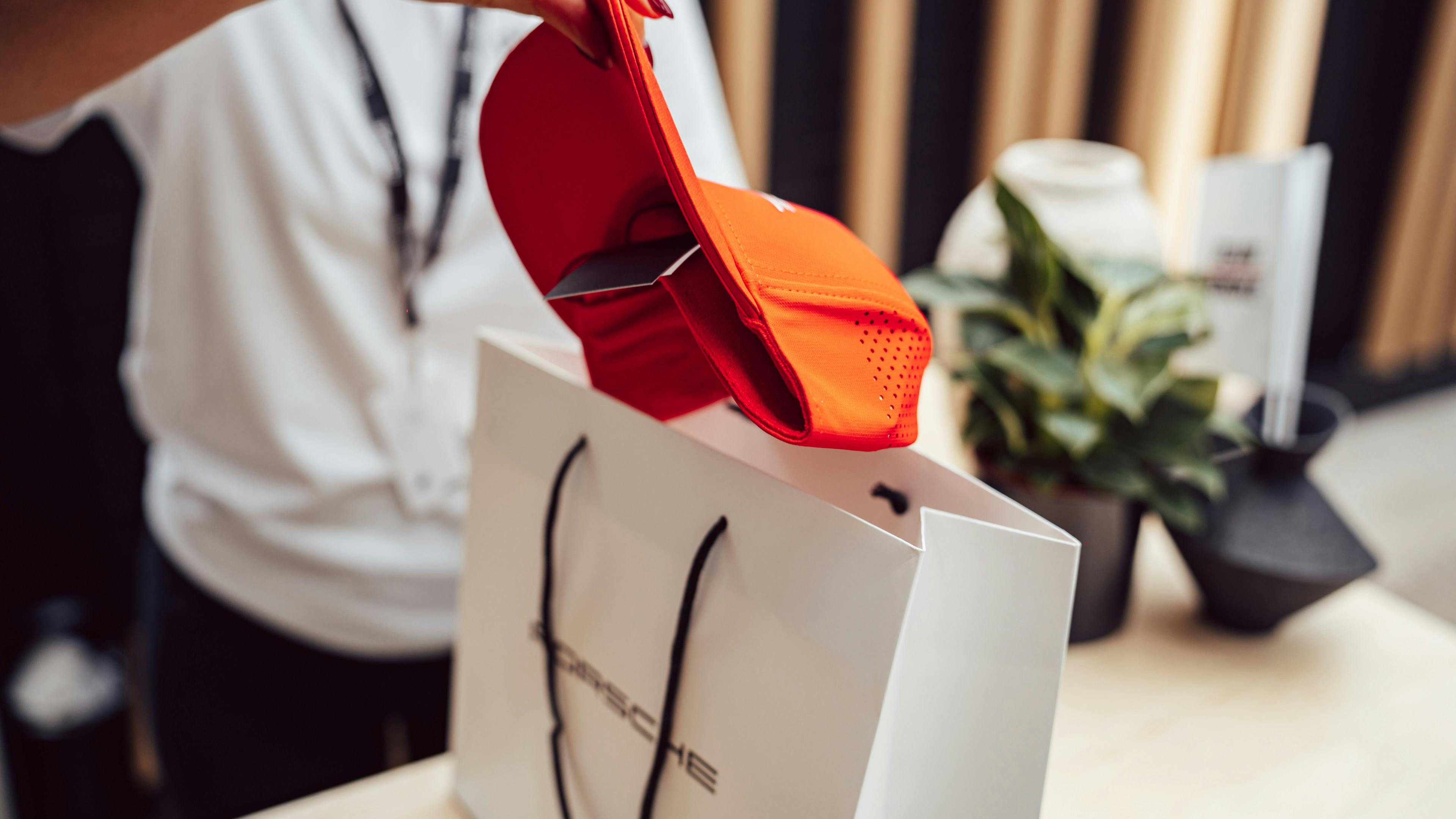 A person placing an orange cap into a white Porsche branded shopping bag on a counter.
