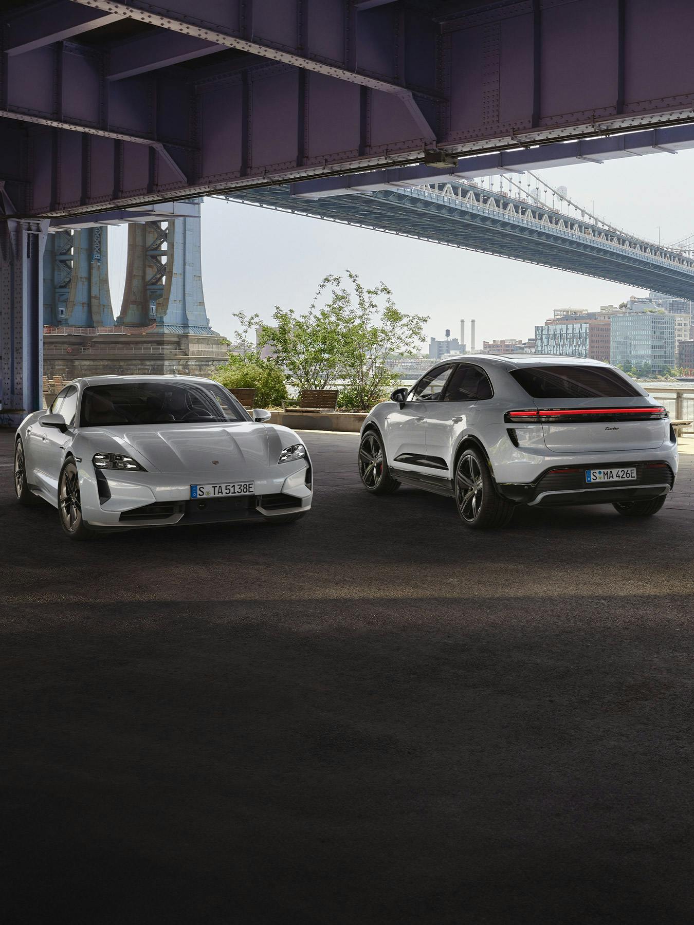 A Taycan and Macan Turbo Electric parked side by side under a large city bridge.