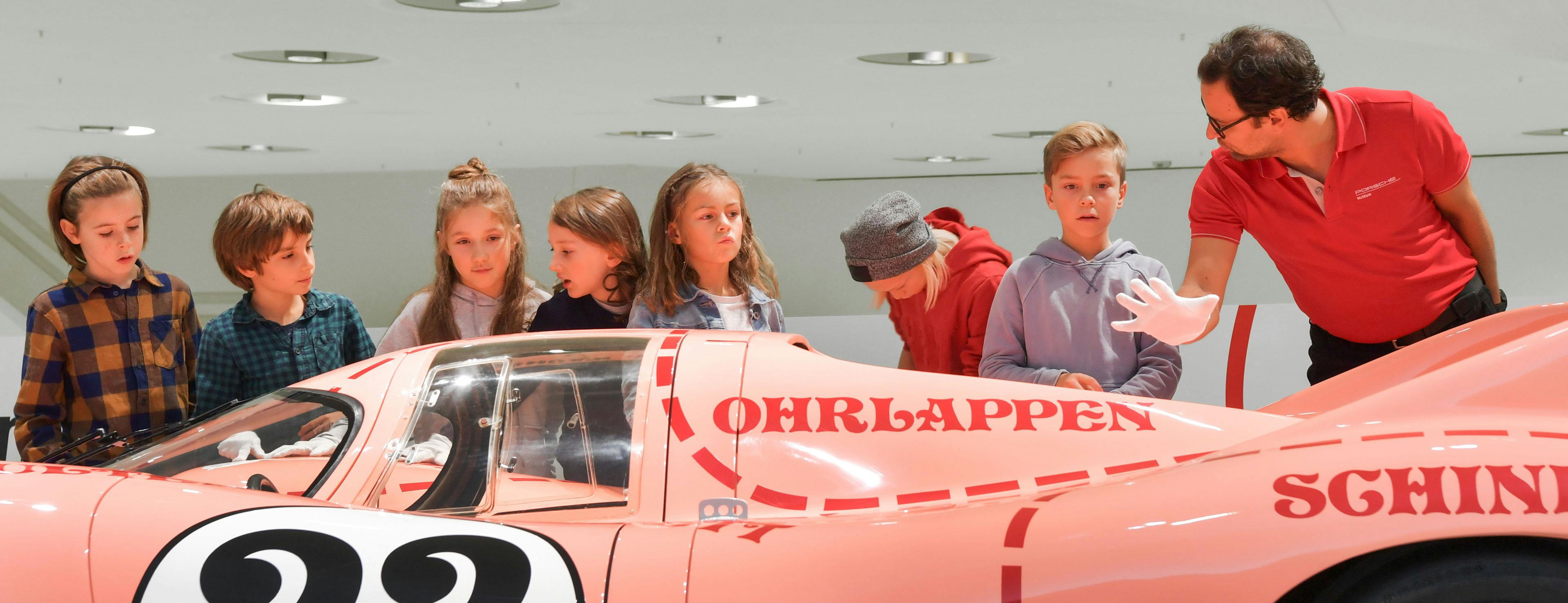Eine Gruppe von Kindern vor einem rosanen Porsche-Exponat mit einem Museumsführer, der über das Auto spricht