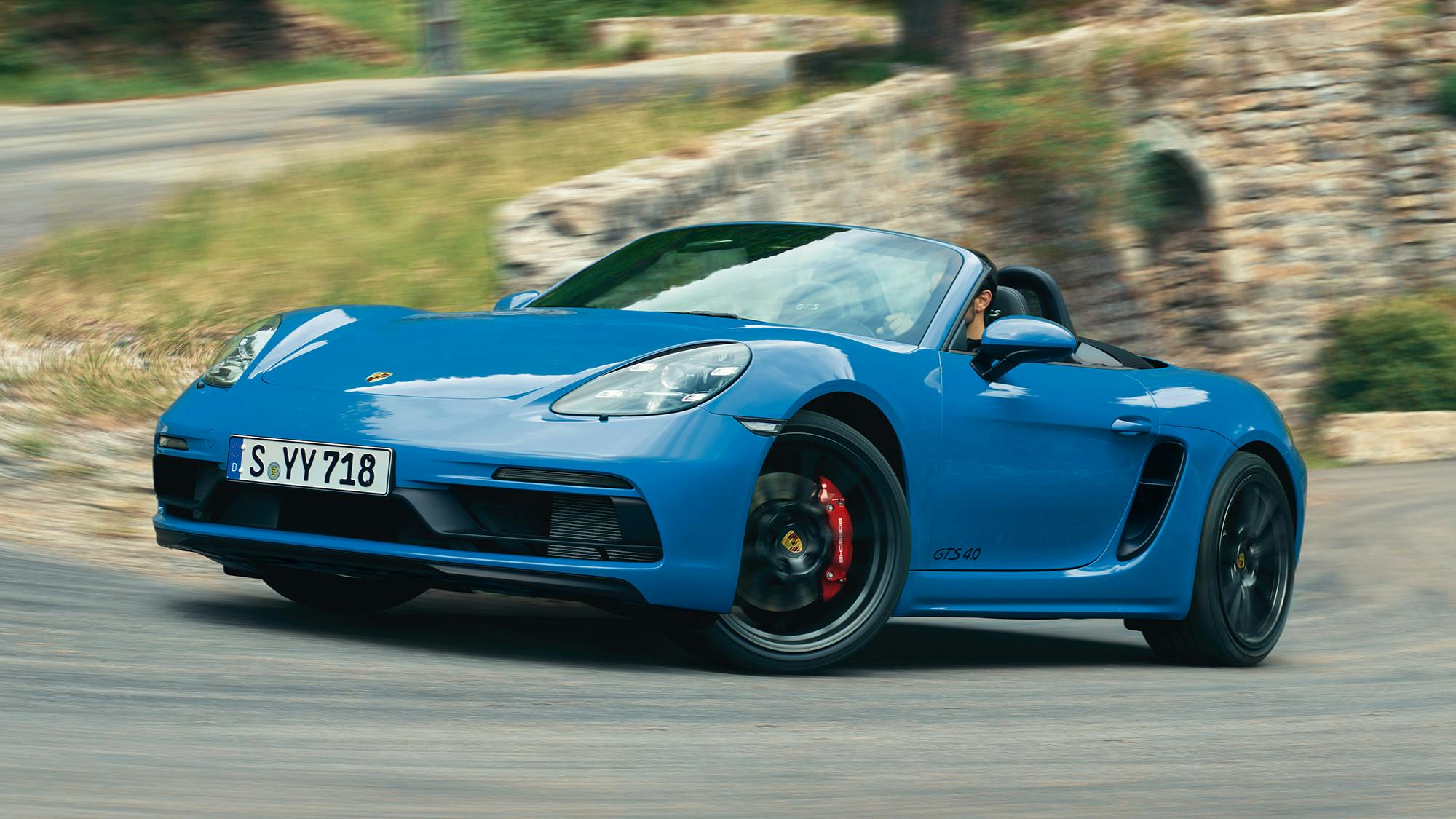 Blauer 718 Boxster fährt schnell durch eine kurvige Bergstraße, umgeben von Bäumen und Felsen.