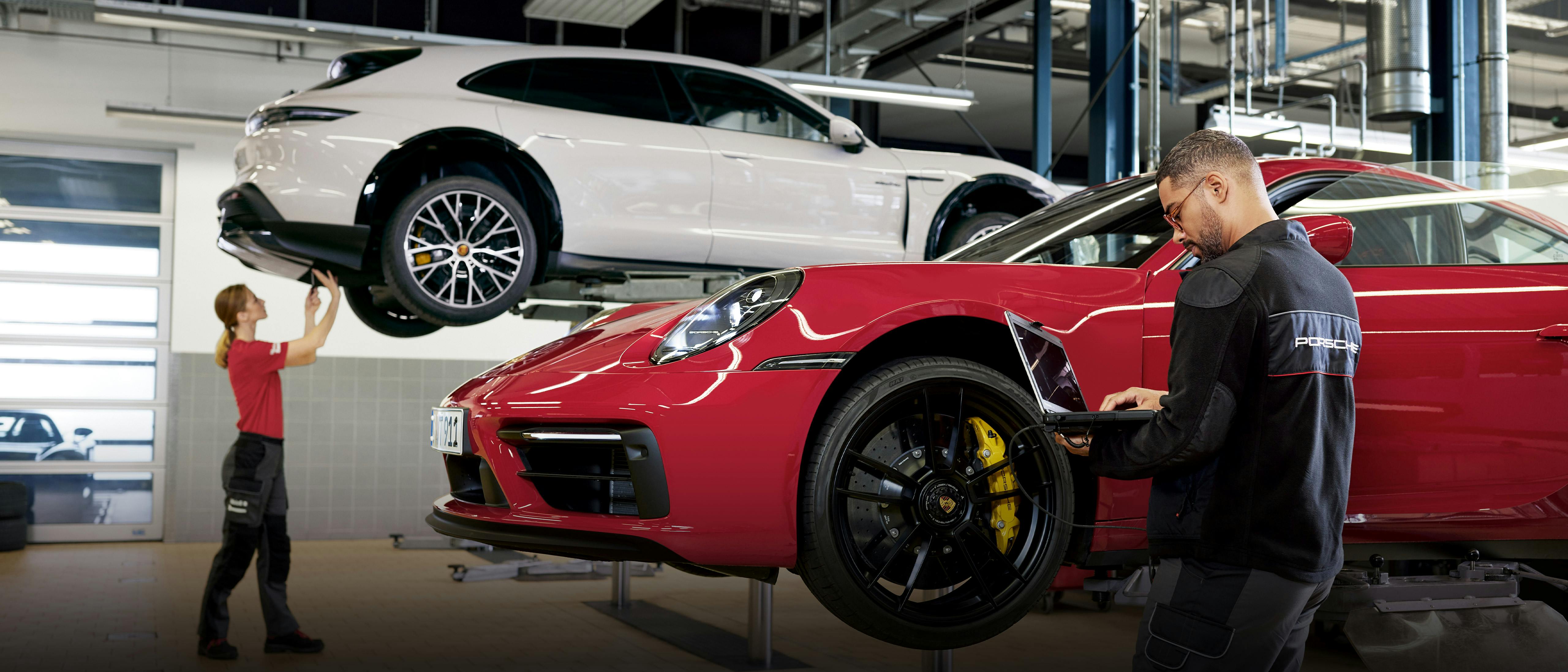 Dans l'atelier Porsche, une employée travaille sur une Porsche blanche et un employé avec un ordinateur portable à côté d'une Porsche rouge, toutes deux sur des plates-formes élévatrices.