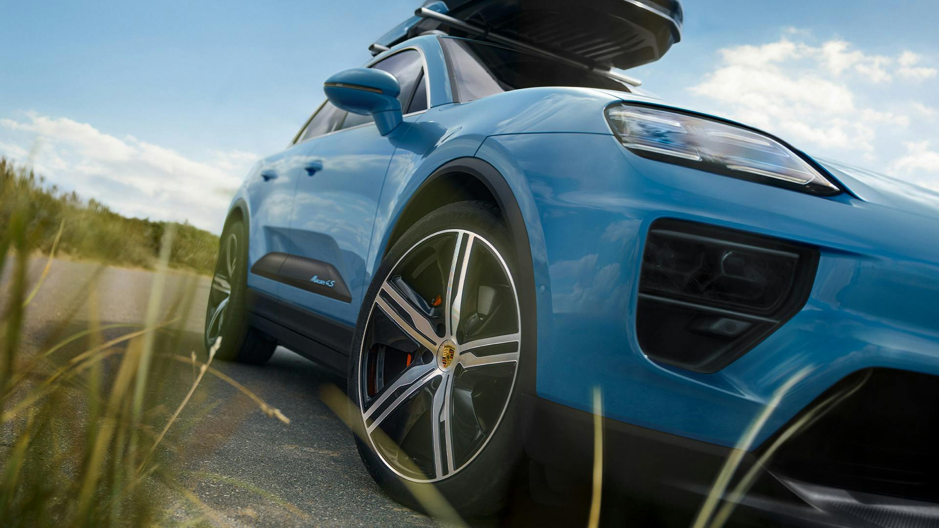 Une Cayenne Turbo E-Hybrid avec cage de toit et planche de surf se trouve à côté d'une 964 Cabrio classique dans les dunes. Un pavillon Porsche est visible à l'arrière-plan.