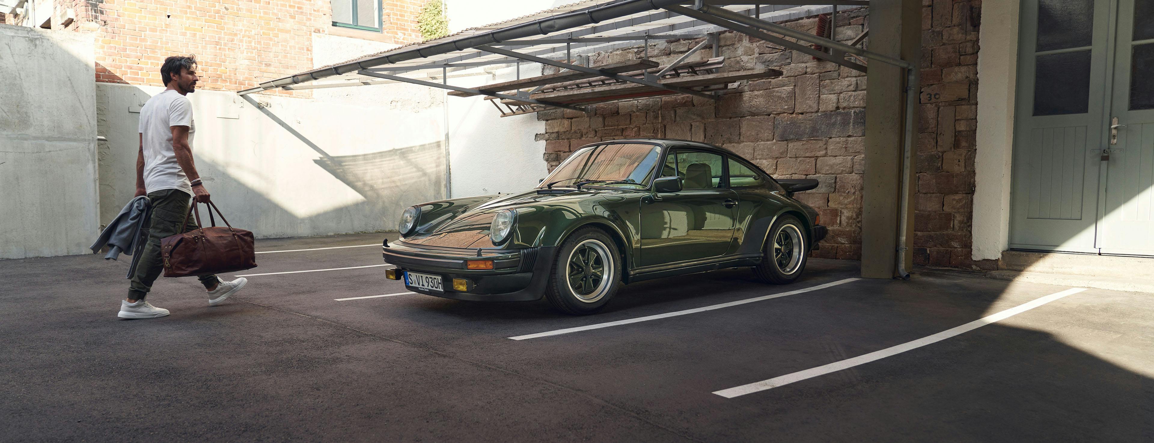 Green Porsche 911 G in a car park, man with sports bag walking towards it.