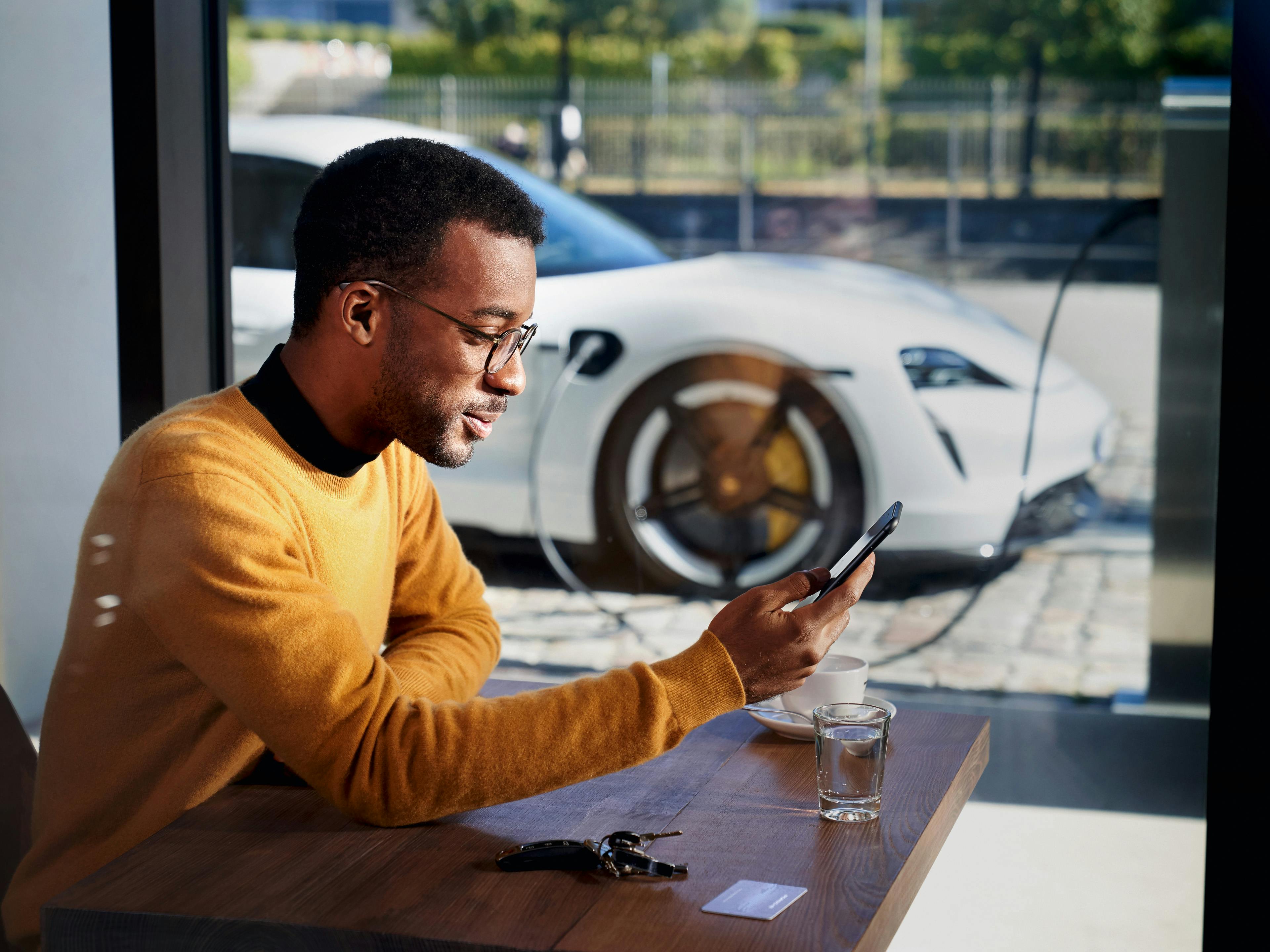 A man in a yellow sweater is looking at his phone, in the background, a white Porsche Taycan is visible.