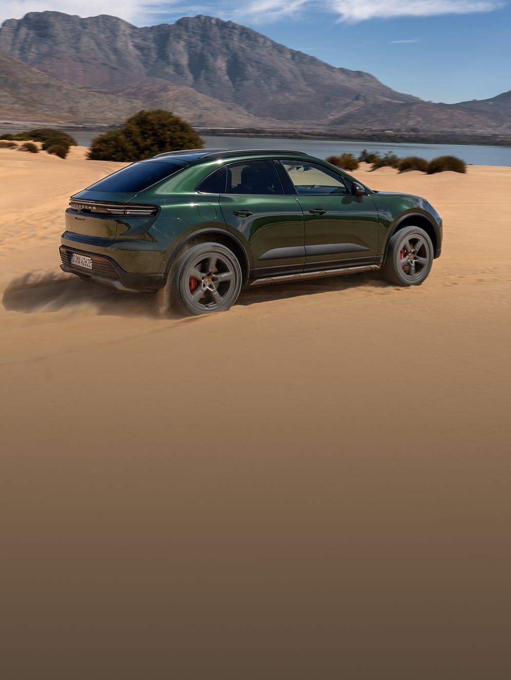 A Porsche Macan 4S driving over sand. A lake and a mountains are in the background.