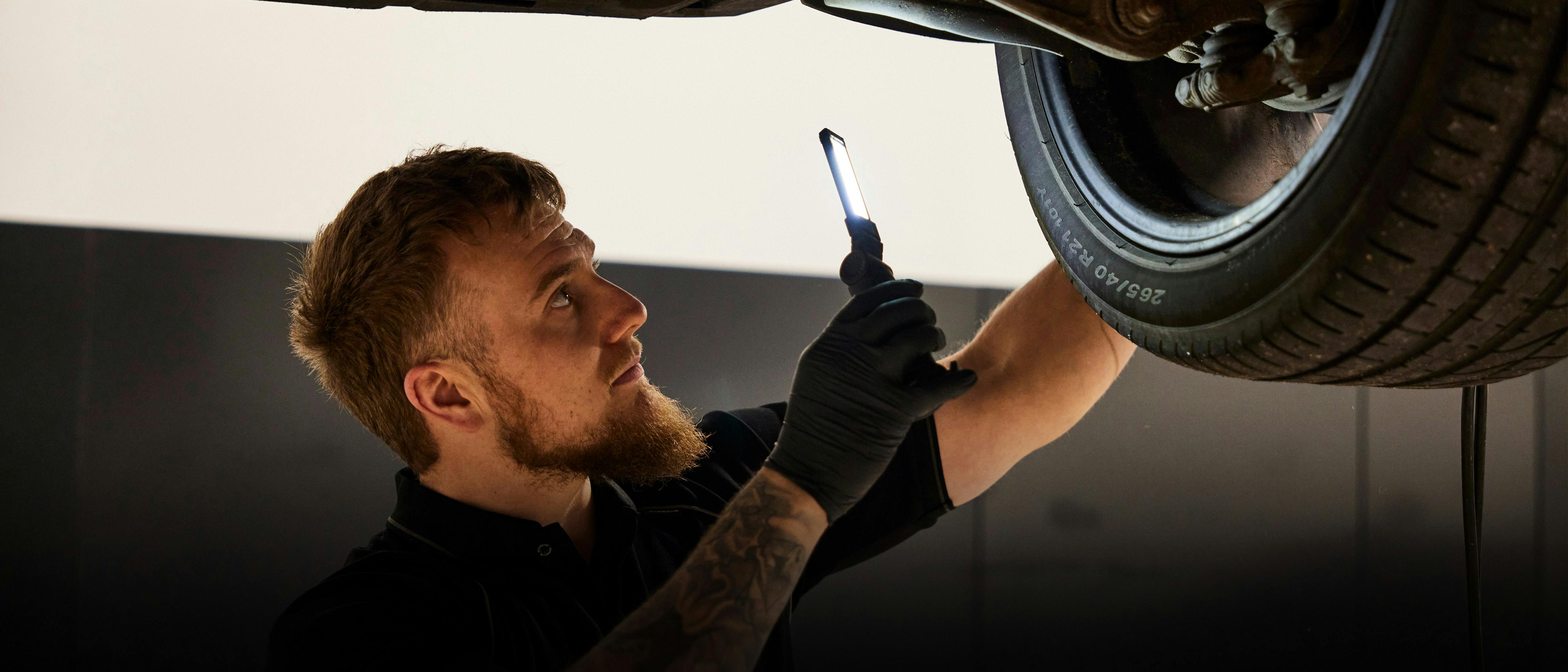 Porsche technician with tattoos inspects suspension and tire of raised vehicle using handheld light.