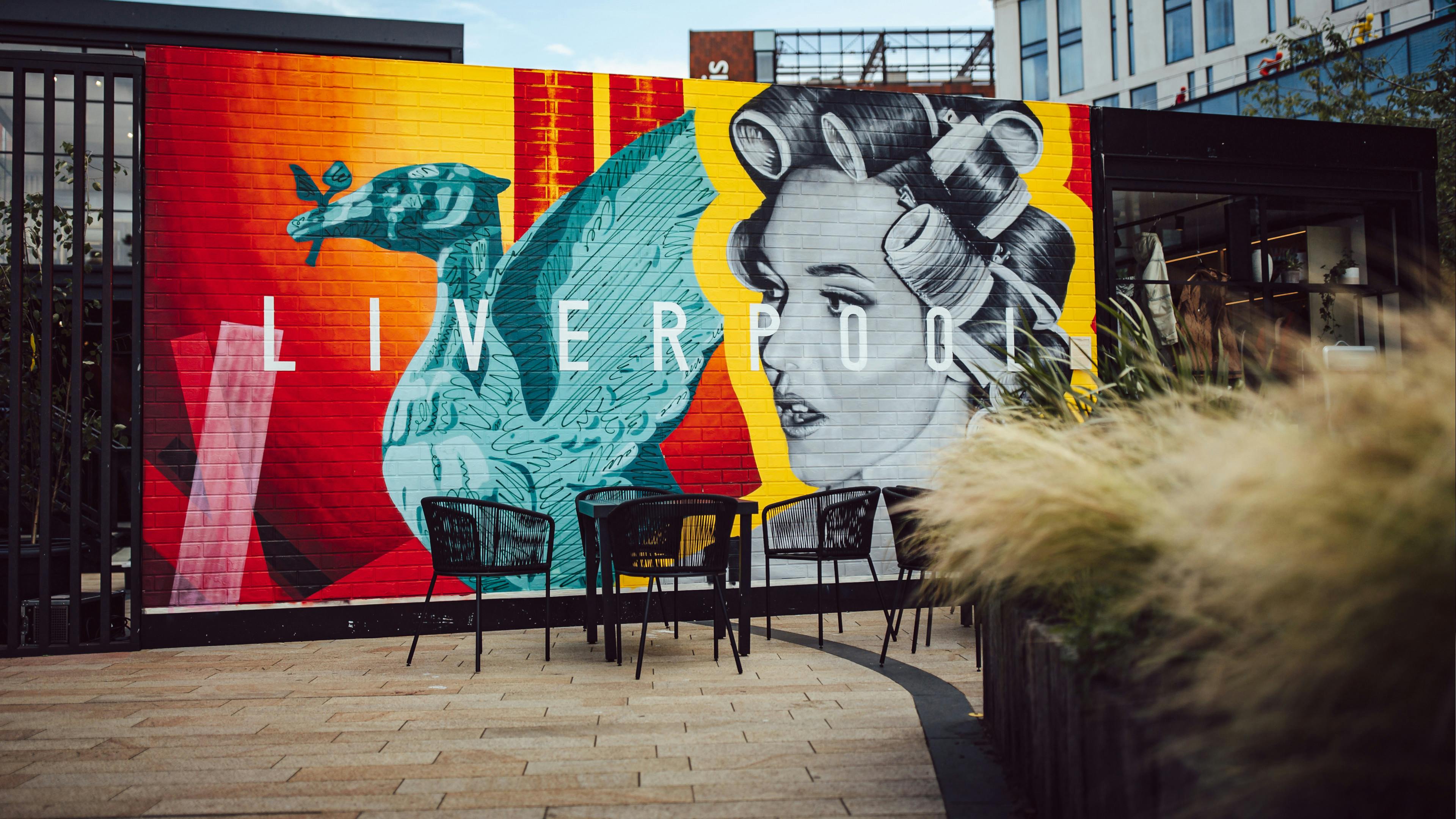 An outdoor patio area featuring a large, vibrant mural on a brick wall that says “LIVERPOOL”. The mural depicts a teal Liver Bird and a black-and-white portrait of a women with her hair in rollers. Several black modern chairs and a table are placed in front of the wall.