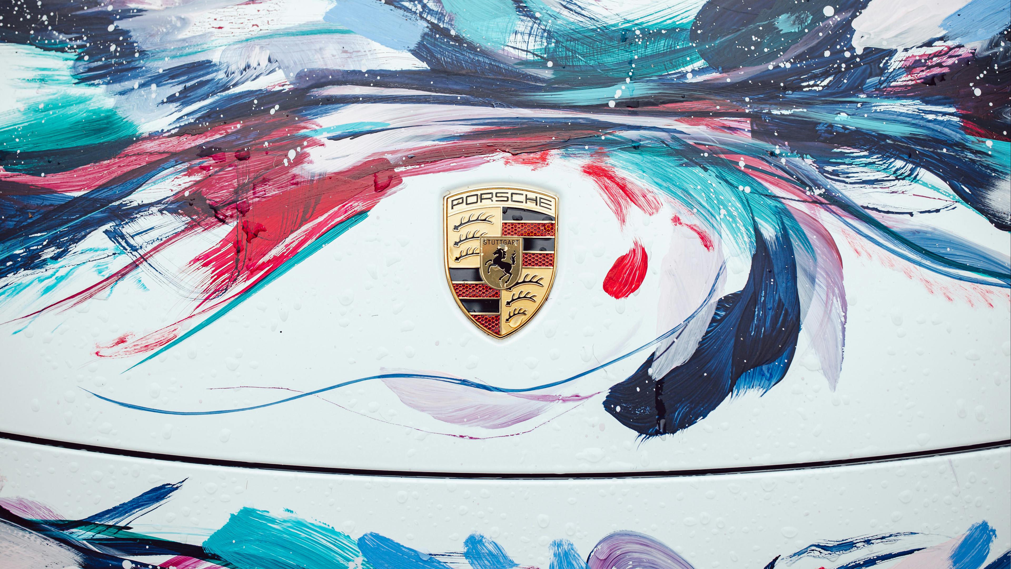 A close-up of the black, red and gold Porsche crest centred on a white car hood. The hood is decorated with expressive, multi-coloured paint strokes in shades of blue, teal, red and purple.