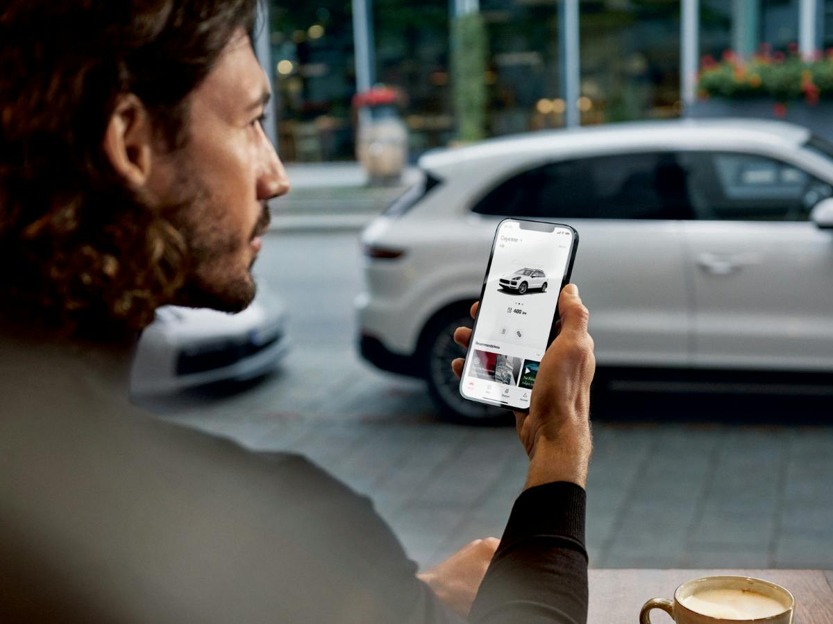 Un homme regarde son téléphone portable sur lequel l'application My Porsche est ouverte. On peut voir une Porsche en arrière-plan.