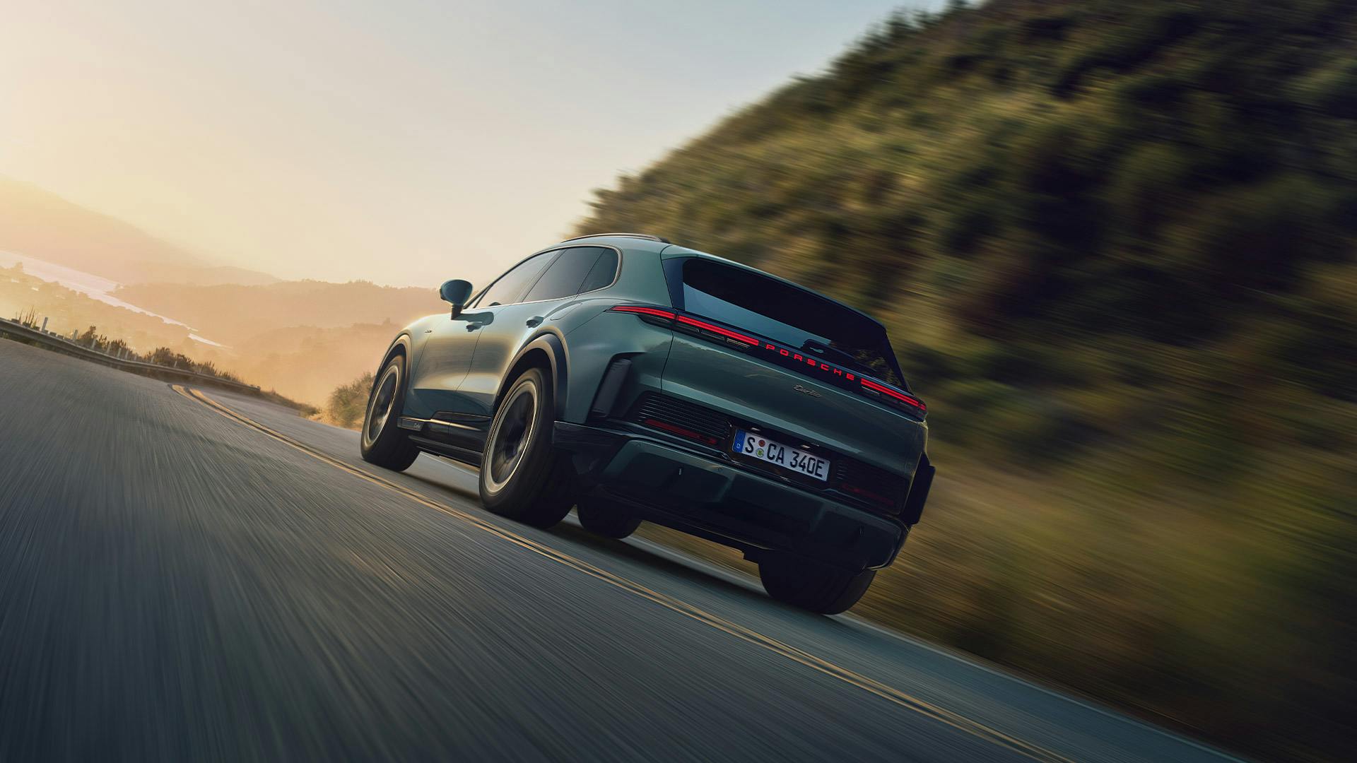 A mystic green metallic Porsche Cayenne Electric drives dynamically along a winding mountain road, surrounded by wooded hills and a sunrise in the background.