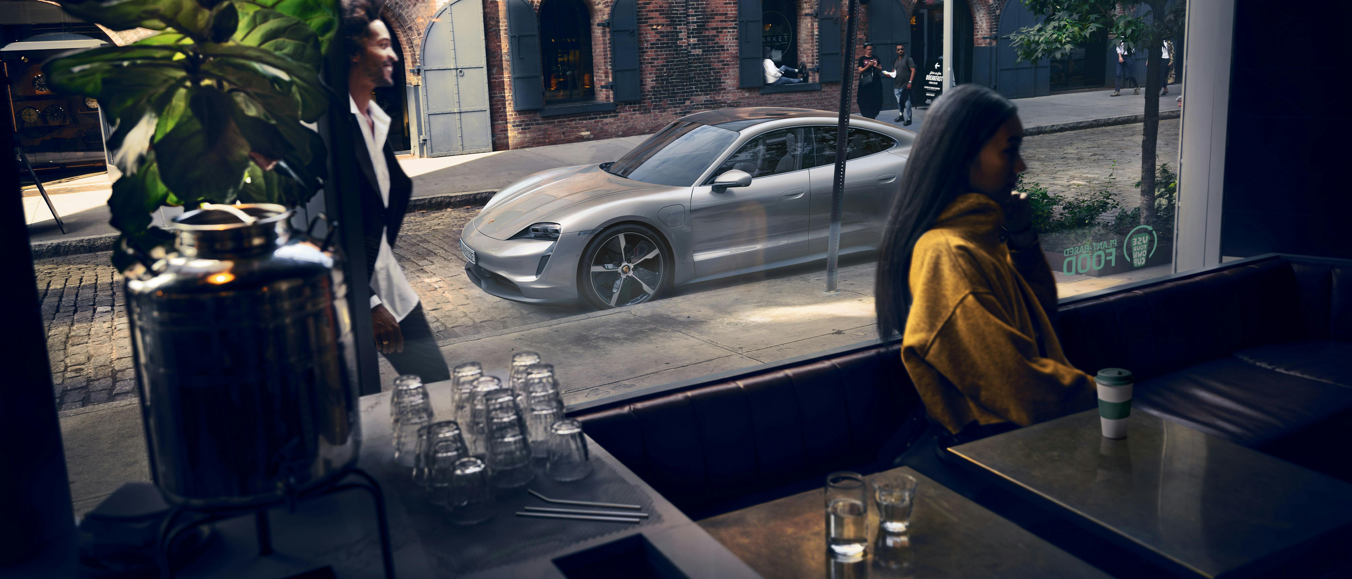 Woman enjoying coffee at a café with her silver Porsche Taycan parked nearby.