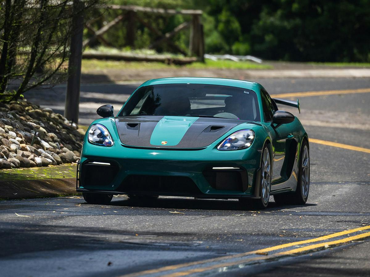 A Porsche 718 GT4 RS in Paint to Sample colour Smyrnagreen is driving on a winding road surrounded by lush green trees and vegetation on a sunny day, shown from the front view.