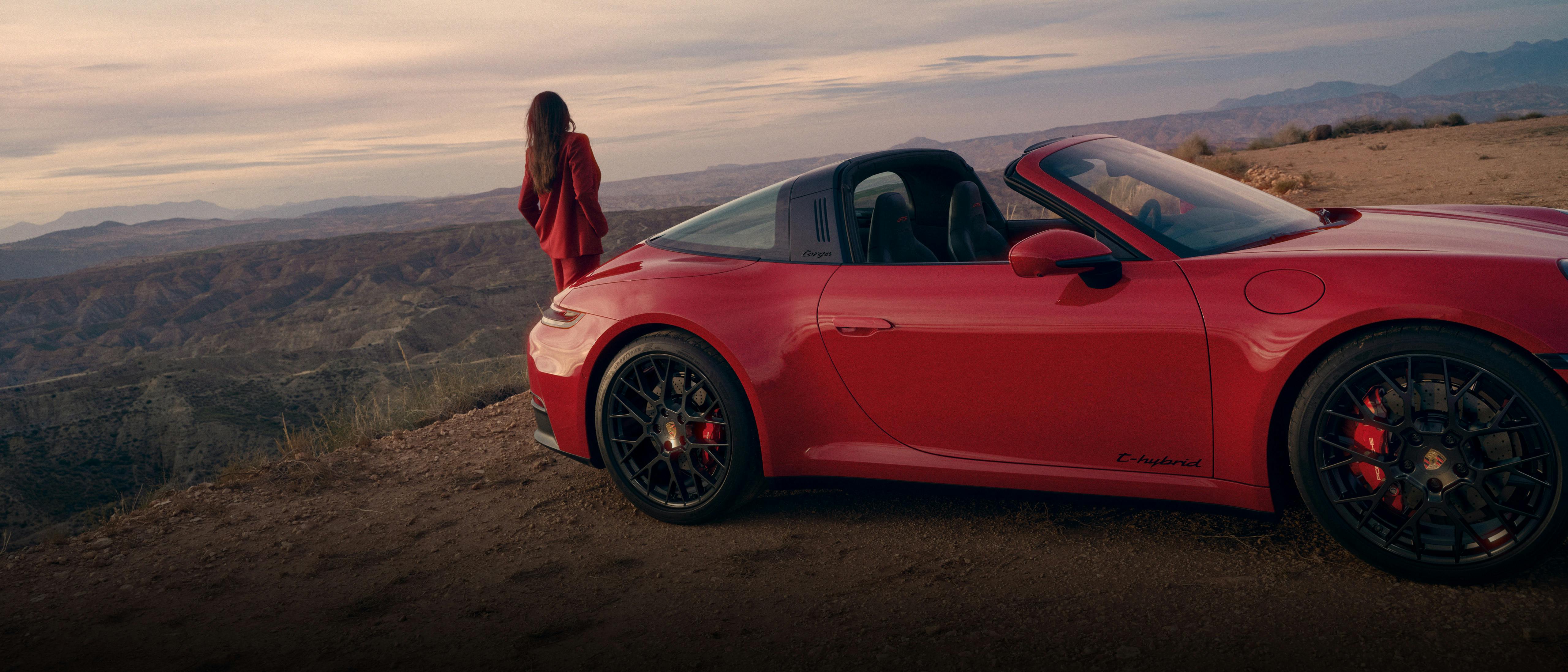 Roter Porsche 911 Targa steht auf einer Anhöhe in einer weiten Landschaft, während eine Person daneben steht und in die Ferne blickt.