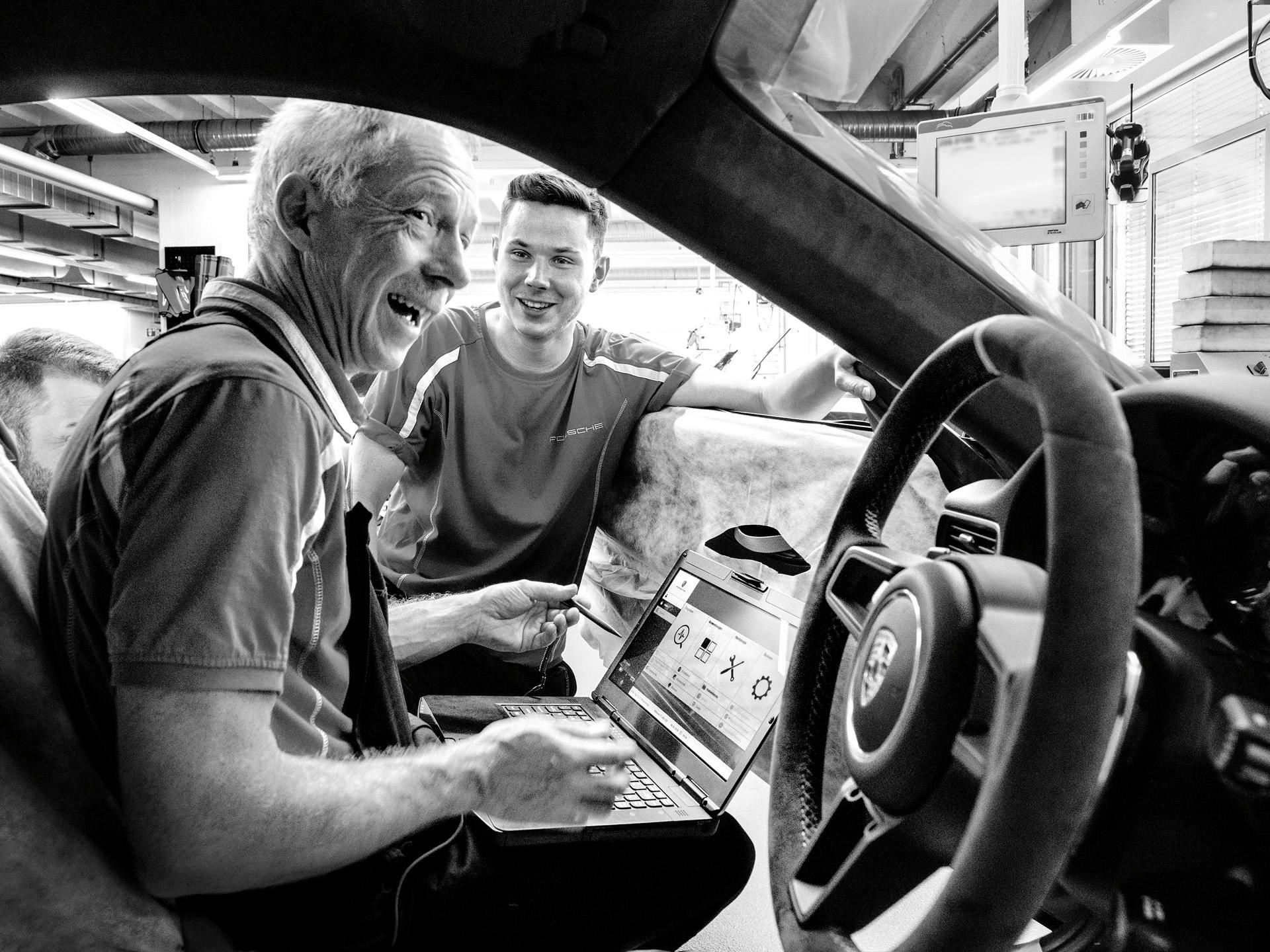 Black-and-white photo: two Porsche employees check the vehicle cockpit on a laptop.