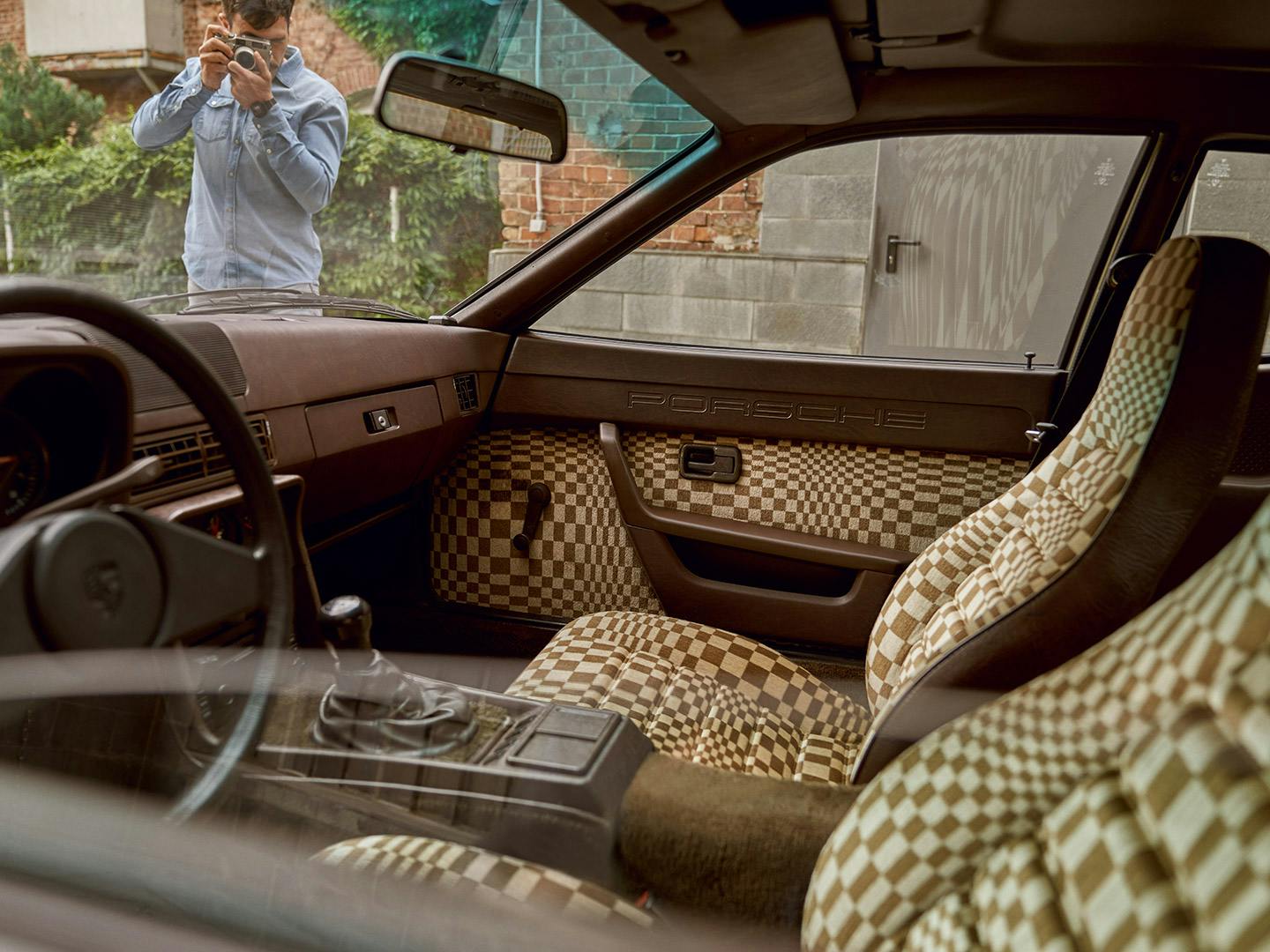 A photographer takes a picture of a classic Porsche vehicle interior featuring a brown and tan 70s-inspiried checkerboard pattern.