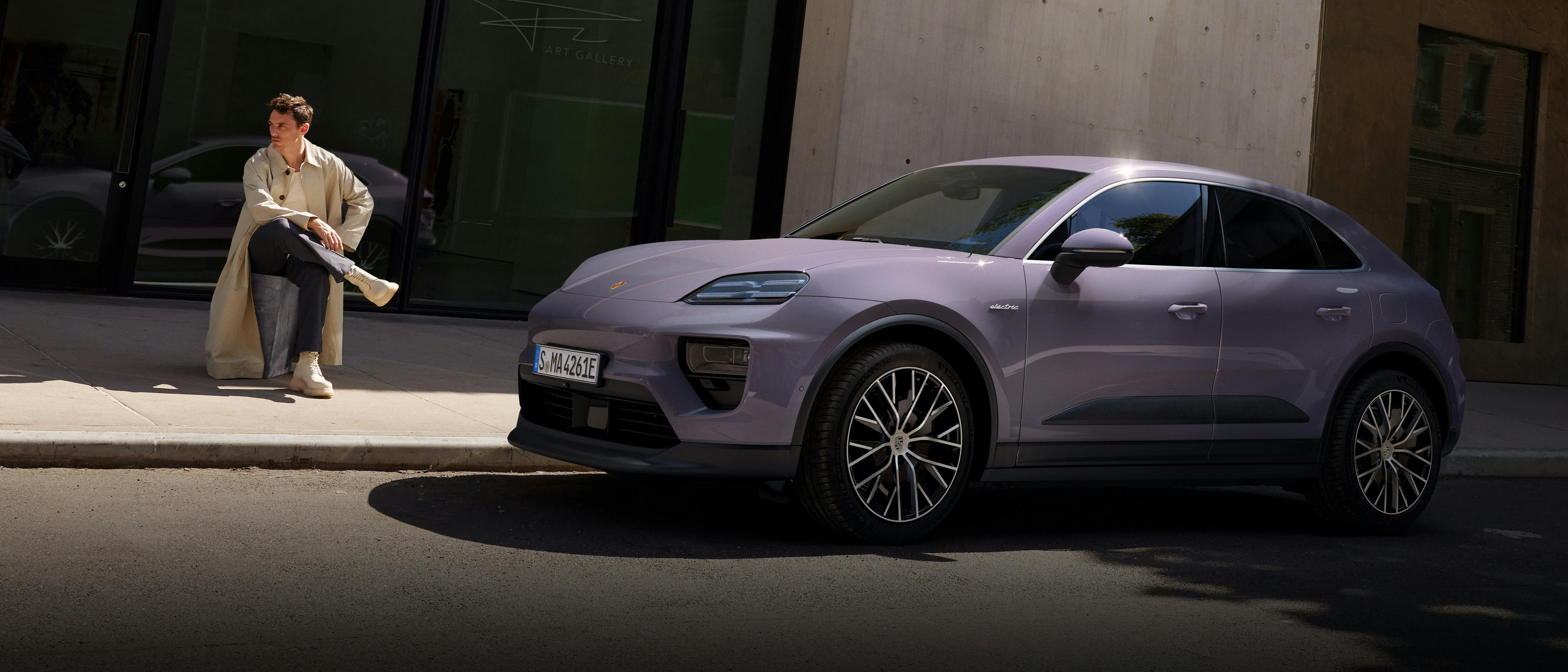 Porsche Macan steht vor einem modernen Betongebäude. Eine Person sitzt links neben dem Fahrzeug auf einer niedrigen Stufe am Gebäude.