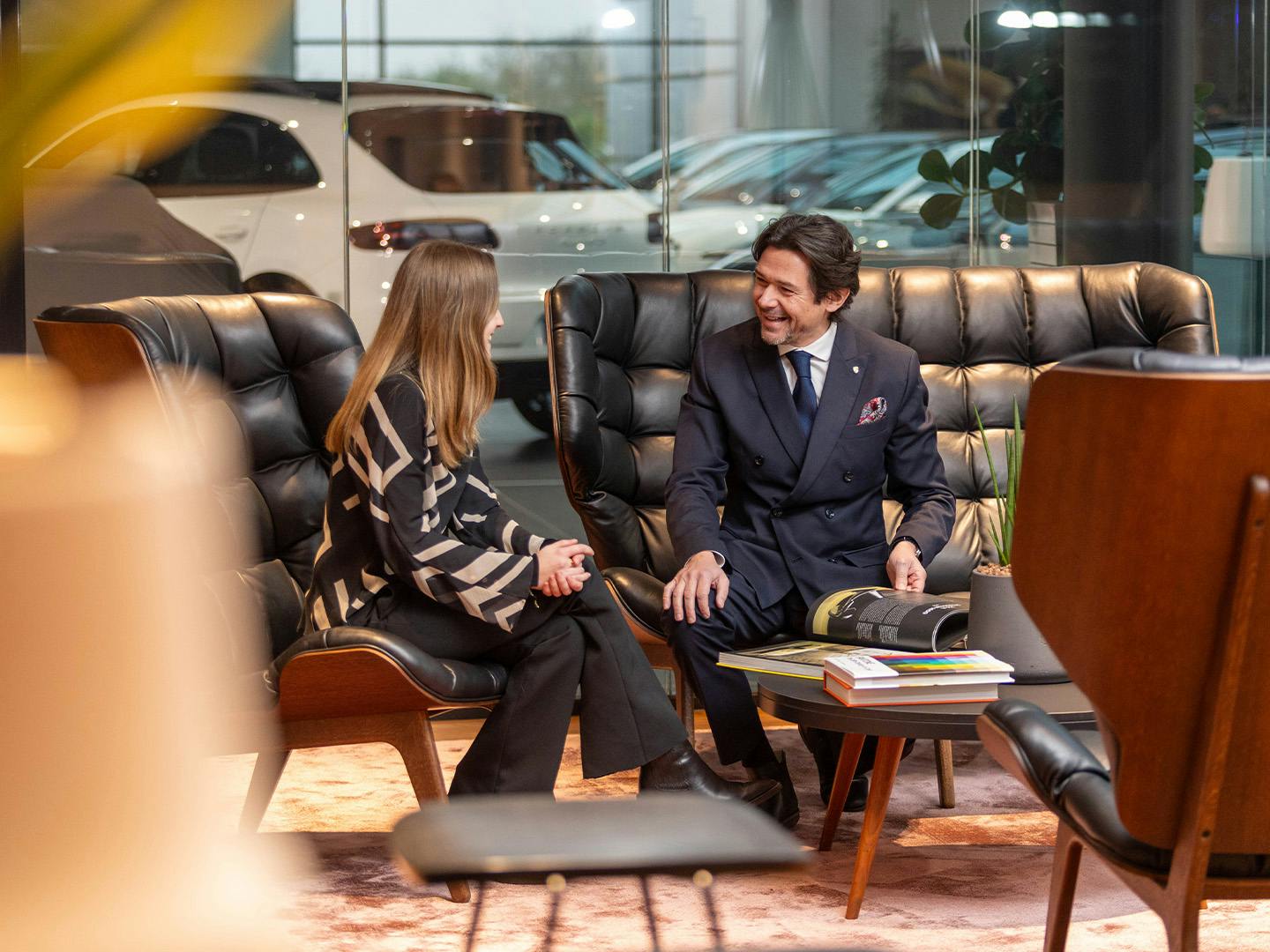 Un employé de Porsche, assis sur un canapé en cuir noir dans un coin salon, parle à une cliente assise dans un fauteuil à côté du canapé.