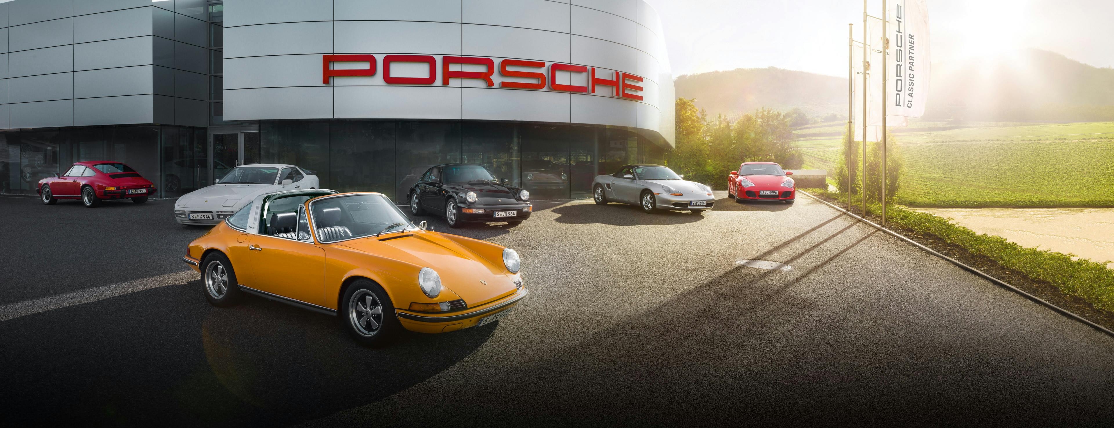 Verschiedene Porsche Modelle (vom 356 bis zum Carrera GT) auf dem Parkplatz eines Classic Partners mit gelbem 911er Carrera Cabriolet Classic im Vordergrund.