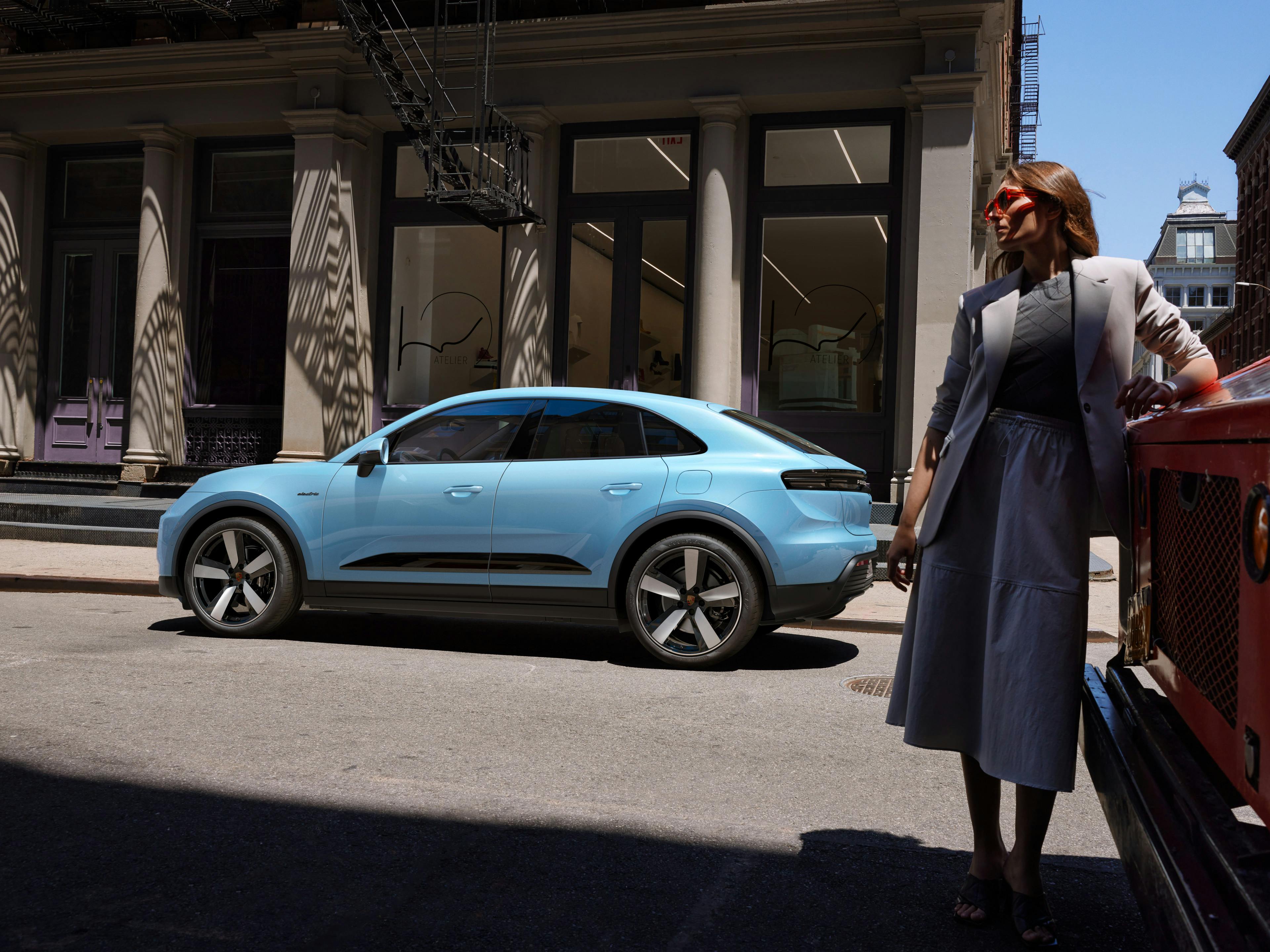 Zwei Porsche Macan Elektro, einer weiß und der andere lila, sind unter einer eisernen Brücke geparkt, mit einer Stadtansicht im Hintergrund.