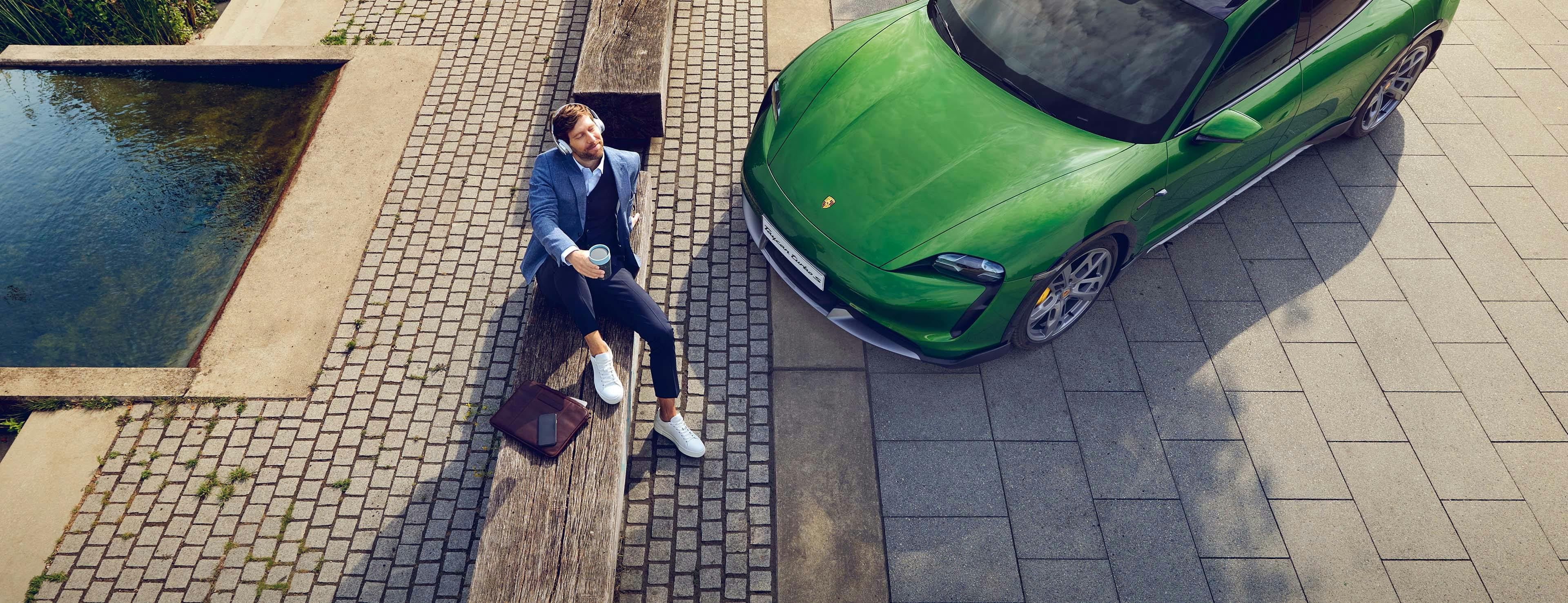 Op een bankje naast een fontein zit een man met een koffie to-go, naast hem staat een groene Porsche geparkeerd.